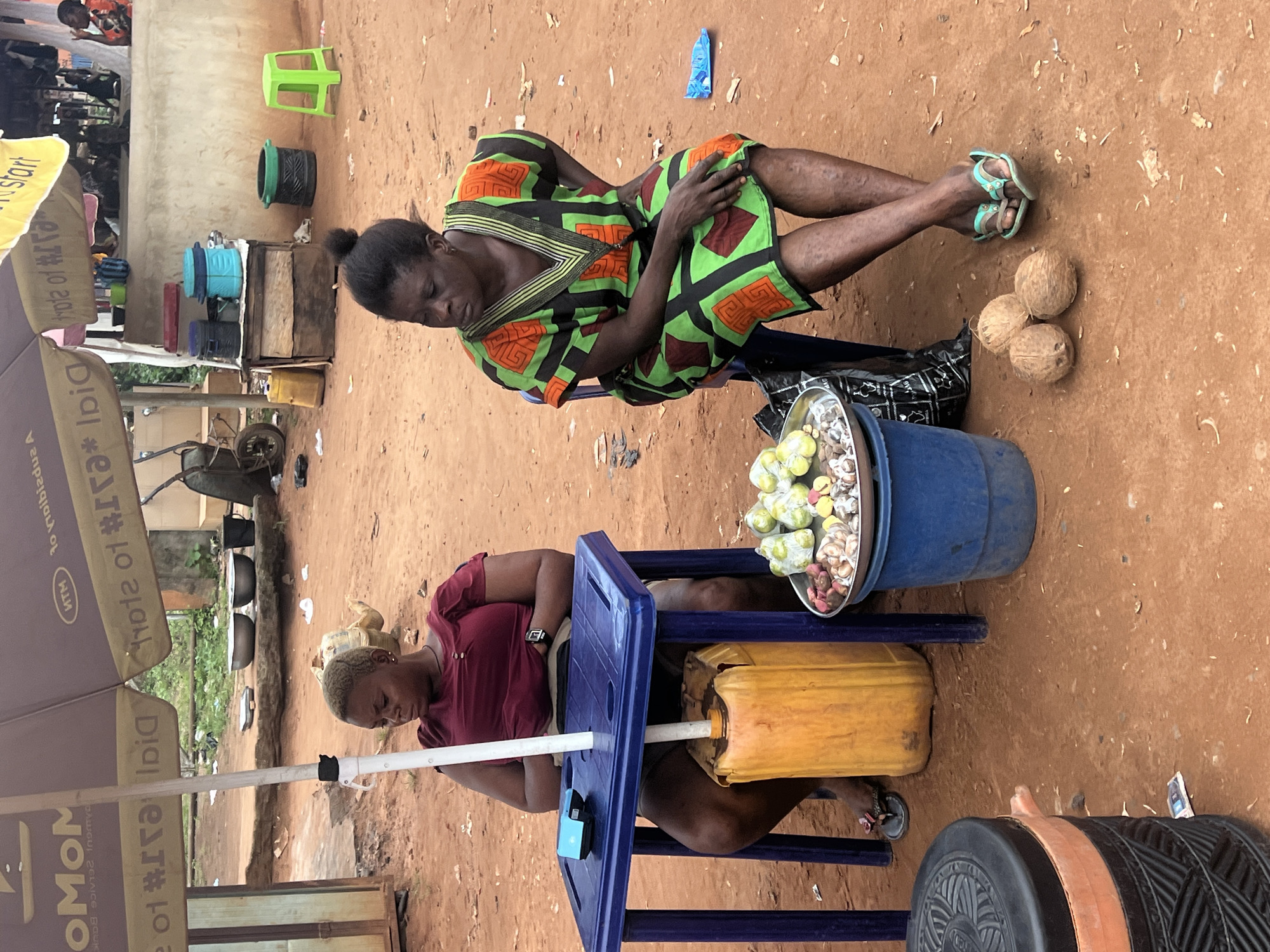 In Nkpor, Nigeria, two women sit by a roadside stall under a large umbrella advertising "MTN" and "Momo". The woman on the left, wearing a maroon shirt, sits behind a small blue table with a yellow plastic container underneath it. On the table, there is a small blue electronic device. The woman on the right, dressed in a green and orange patterned dress, sits on a chair. In front of her, on the ground, are three coconuts. To her left, a blue bucket holds a tray of produce, including green fruits and nuts. The ground is covered in reddish-brown dirt, with various objects scattered around, including plastic bags and debris. In the background, a cart, metal bowls, and colorful plastic containers are visible, suggesting a busy market environment.