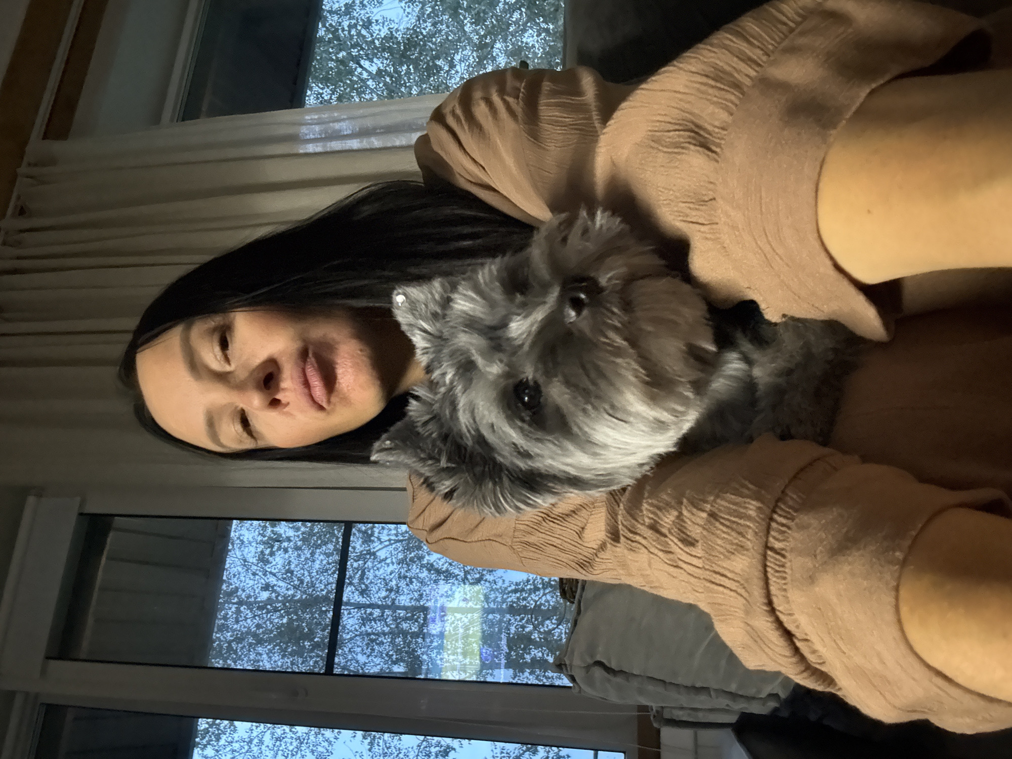 The image shows a woman holding a small, grey dog. The woman has long, dark hair and is wearing a brown, textured top. Her face is visible, and she appears to be looking at the camera with a neutral expression. The dog is held close to her chest, with its head resting on her shoulder. The dog has a scruffy coat and dark eyes.
The scene appears to be indoors, likely in a home. Through a window in the background, trees with bare branches are visible against a twilight sky, suggesting the image was taken during the evening. The lighting is dim, contributing to a cozy atmosphere. The overall mood conveyed is one of affection and companionship between the woman and her dog. There is no visible text in the image.