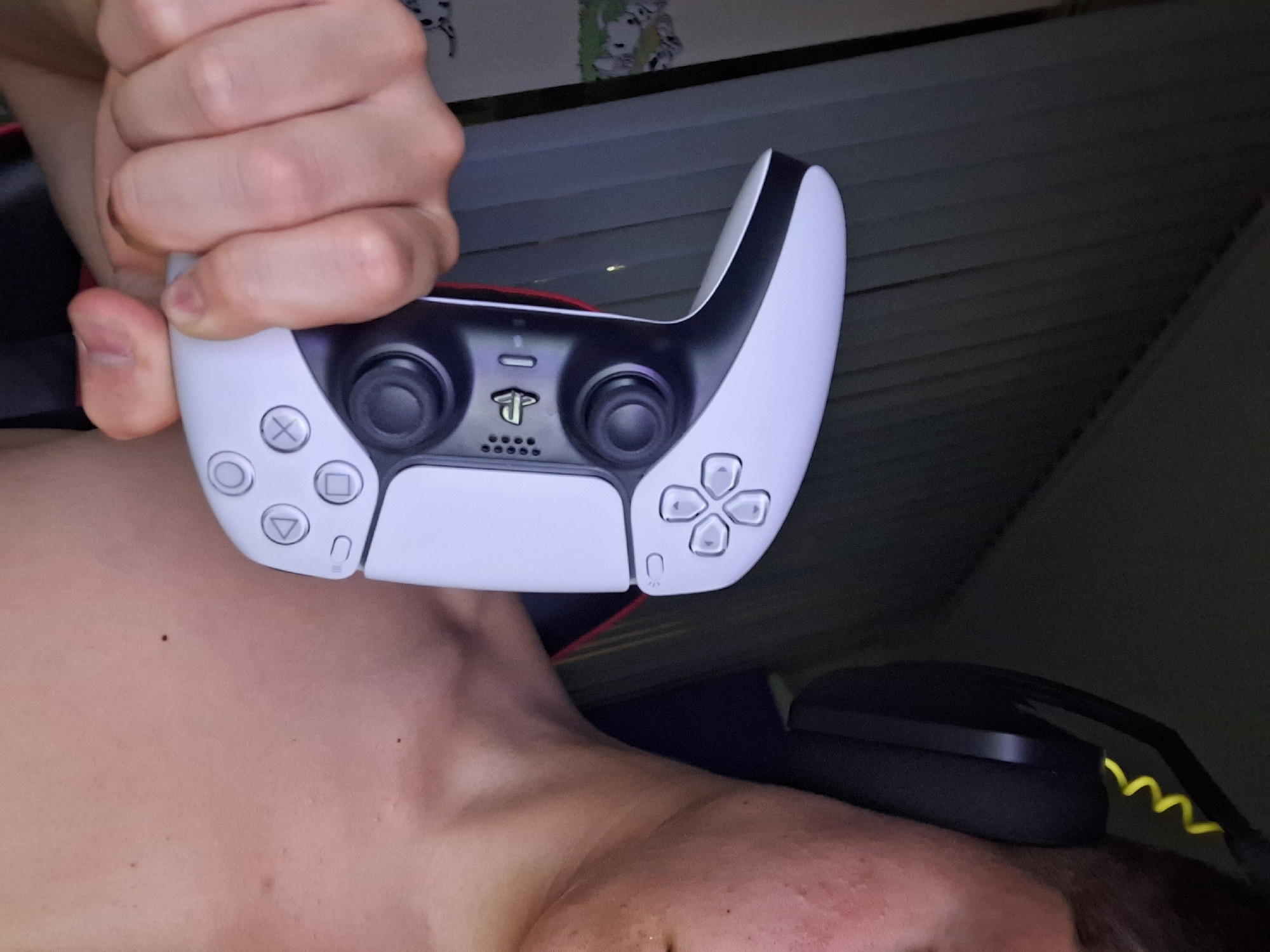 A person's bare shoulder and neck, with some minor skin blemishes, are visible on the right side of the frame, partially obscured by a black gaming headset with a yellow coiled wire. The person is holding a white and black PlayStation 5 controller in their left hand, which is in the foreground. The controller has its directional buttons and analog stick visible. The background consists of dark curtains and a partially visible wall with a framed image of cartoon dogs. The scene appears to be indoors, likely in a residential setting, and there is no indication of the city of Varna, Bulgaria.