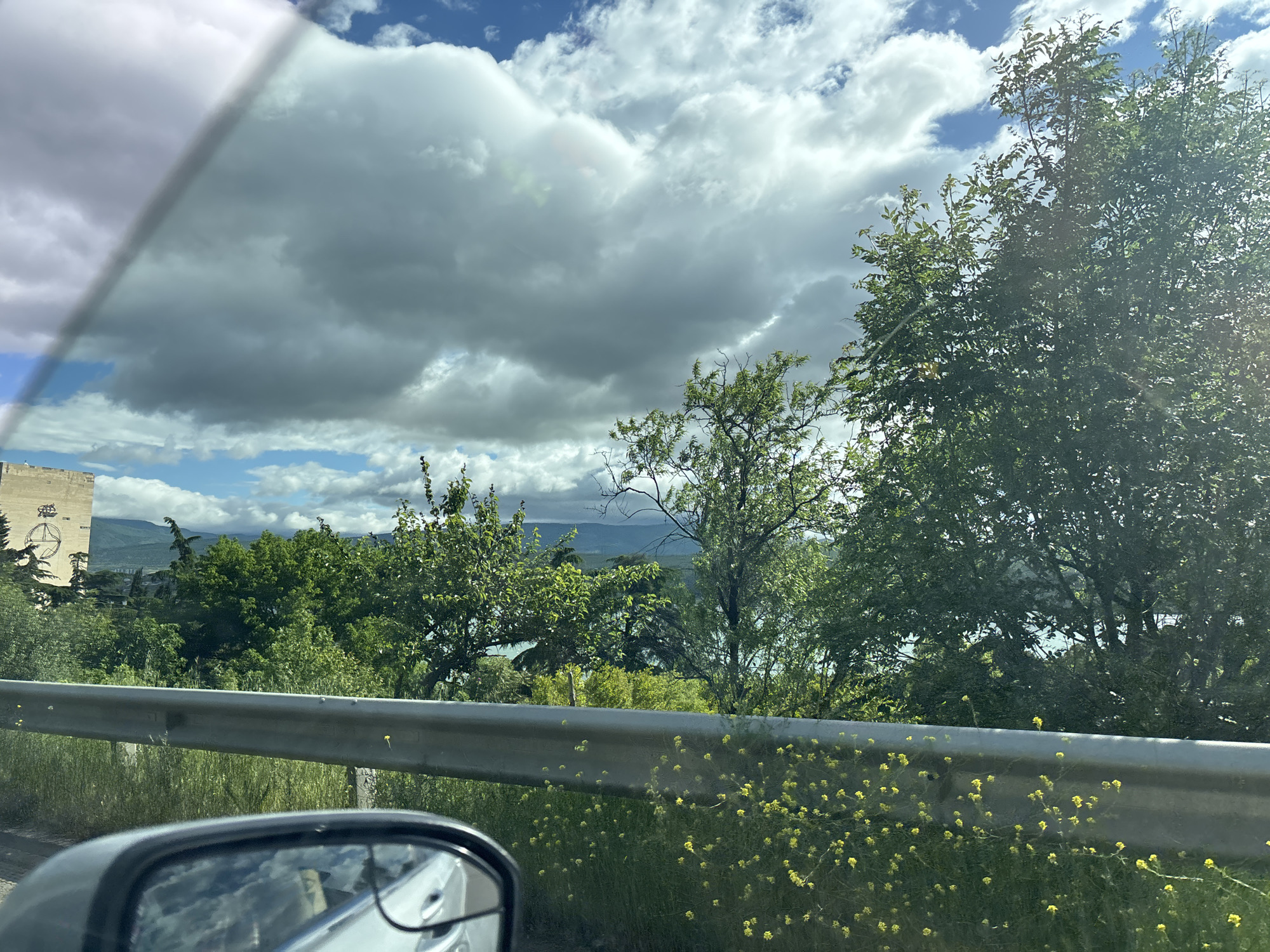 The image, taken from inside a car, shows a landscape with a mix of natural and man-made elements. The foreground features a gray guardrail with small yellow flowers growing near it. Behind the guardrail is dense greenery comprised of trees and bushes. A tall, rectangular structure, possibly a building or tower, can be seen in the distance, marked with what appear to be drawings or symbols. This structure is located in Tbilisi, Georgia. The sky is filled with white and gray clouds, with patches of blue visible. The car's side mirror is visible in the lower left corner.