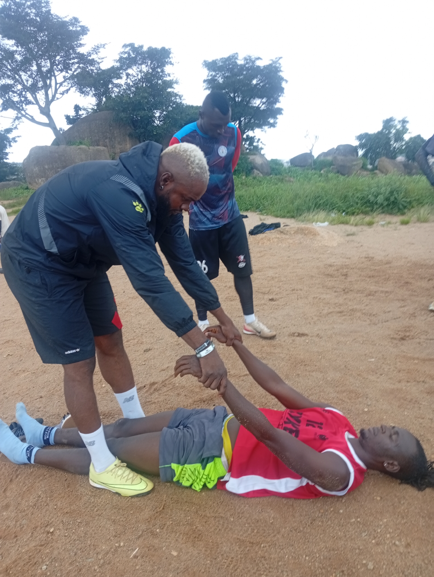 In Jos, Nigeria, a group of individuals appear to be engaged in physical training. One person, lying on their back in the foreground, is wearing a red tank top and grey shorts with yellow trim. Their arms are extended upwards and appear to be held by another person standing over them. This standing individual, in the mid-ground, is wearing a dark jacket and shorts, with blonde-tipped hair. Another person, in the background, is wearing a blue and red patterned athletic shirt and dark shorts. The setting is an outdoor, sandy area with sparse vegetation and large rocks in the background, indicative of a training ground or natural landscape.