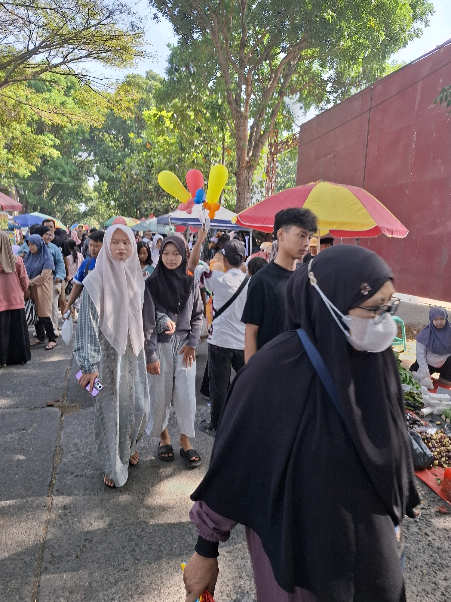 This is a street-level, eye-level, medium shot photograph taken in the city of Karangtengah, Indonesia, showing a bustling outdoor market scene. The foreground is dominated by the back of a person wearing a black hijab and a purple garment underneath, obscuring most of their body. This individual is holding what appears to be a part of a colorful umbrella or toy. To the left of this person, two young women are walking towards the camera. The woman on the left wears a pale pink hijab and a patterned grey dress, and she is holding a purple mobile phone. The woman to her right has a black hijab and is wearing grey pants and a grey hooded top.
Behind these individuals, the market is crowded with people of various ages, many of whom are wearing hijabs and modest clothing. Stalls are set up along the street, indicated by colorful umbrellas and awnings. In the mid-ground, a young man in a black t-shirt is visible, looking to his left. Further into the background, more people are walking and browsing, and market stalls are abundant.
The environment appears to be a street or pathway, paved with asphalt. Trees with lush green foliage line the sides of the pathway, casting shadows. In the background on the right, a large, plain red wall is visible. The lighting suggests it is daytime, possibly late morning or early afternoon, with sunlight filtering through the trees. Several colorful balloons, red, yellow, and blue, are visible floating above the crowd in the mid-ground. The overall atmosphere is one of a lively community gathering or event.