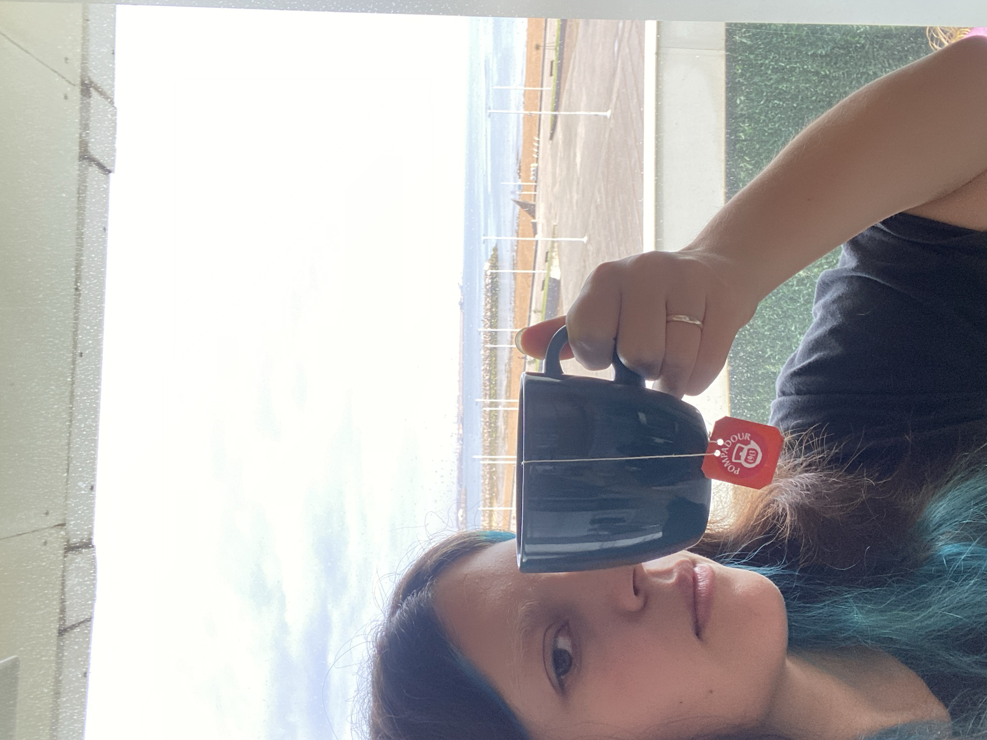 A person is holding a dark blue mug with a tea bag in front of their face, indoors near a window. The tea bag tag is red with white writing. The person has brown hair with streaks of blue and is wearing a dark grey top. Outside the window, a paved area, possibly a promenade or parking area, runs along the coastline of Gijón/Xixón, Spain. There are regularly spaced white posts along the promenade. The sea and the sky are visible in the background, with clouds suggesting an overcast day. Part of a building, possibly a balcony or window frame, is visible to the left, with green foliage visible on the right. The mug appears to be held at roughly eye level.