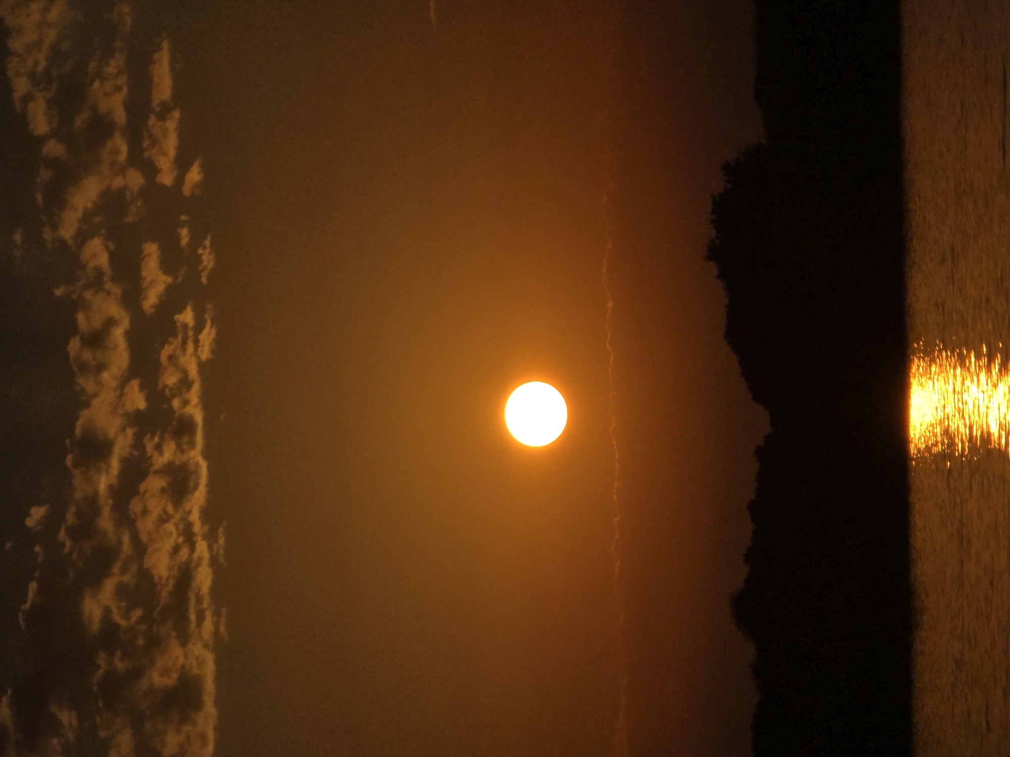 A sunset over the Black Sea at Chernomorets, Bulgaria. The sun is a bright, round orb in the center of the frame, casting a warm orange glow. Clouds are scattered across the sky, tinged with the same orange hue. Below the sky, a dark silhouette of trees forms the horizon. The water in the foreground reflects the sun's light, creating a shimmering path across the surface. The scene is serene and picturesque, capturing the beauty of a coastal sunset.