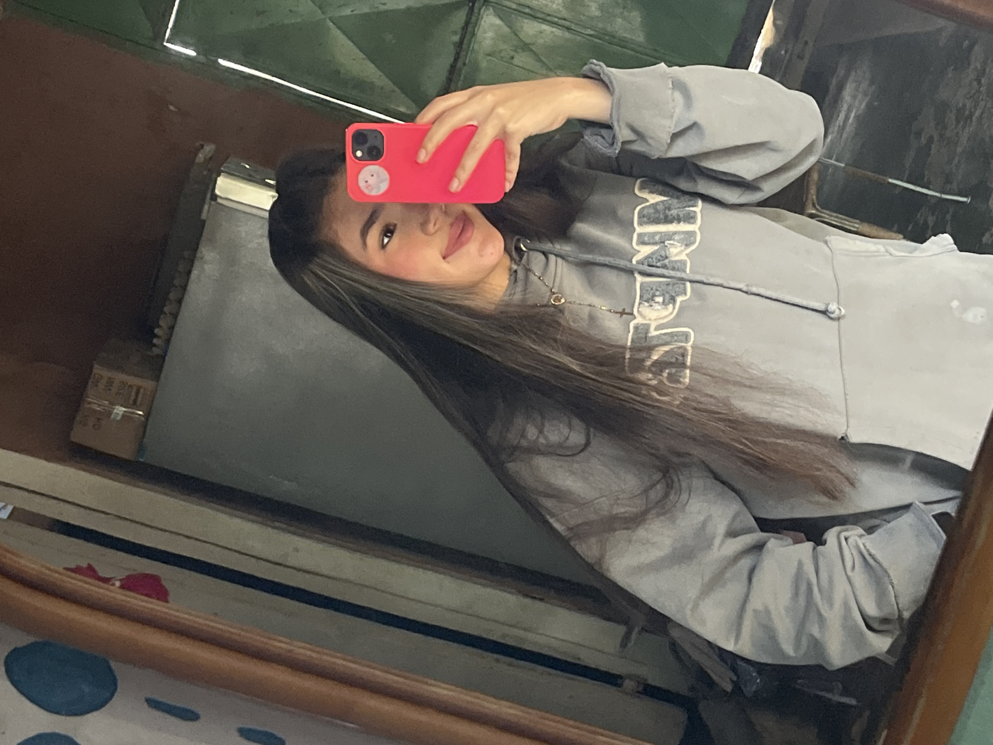 The media file shows a young person taking a mirror selfie. The person is wearing a grey hoodie with a subtle graphic on the chest and a delicate necklace. They are holding a bright pink phone in their right hand, which is angled to capture the reflection. Their left arm is bent, and their hand is resting on the edge of what appears to be a wooden frame, likely part of the mirror. The person has long, dark hair with lighter streaks, and they are smiling softly at the camera. The reflection reveals a dimly lit, somewhat cluttered indoor environment. Behind the person, there's a glimpse of a green, textured surface that might be a wall or ceiling. To the left of the person, a cardboard box is visible, and the overall setting suggests a casual, personal space. The lighting appears to be artificial, with bright reflections suggesting overhead lights or possibly sunlight filtering in from a window. The image has a slightly desaturated and warm tone. There is no visible text in the media file. The context provided indicates the location is Juan Griego, Venezuela.