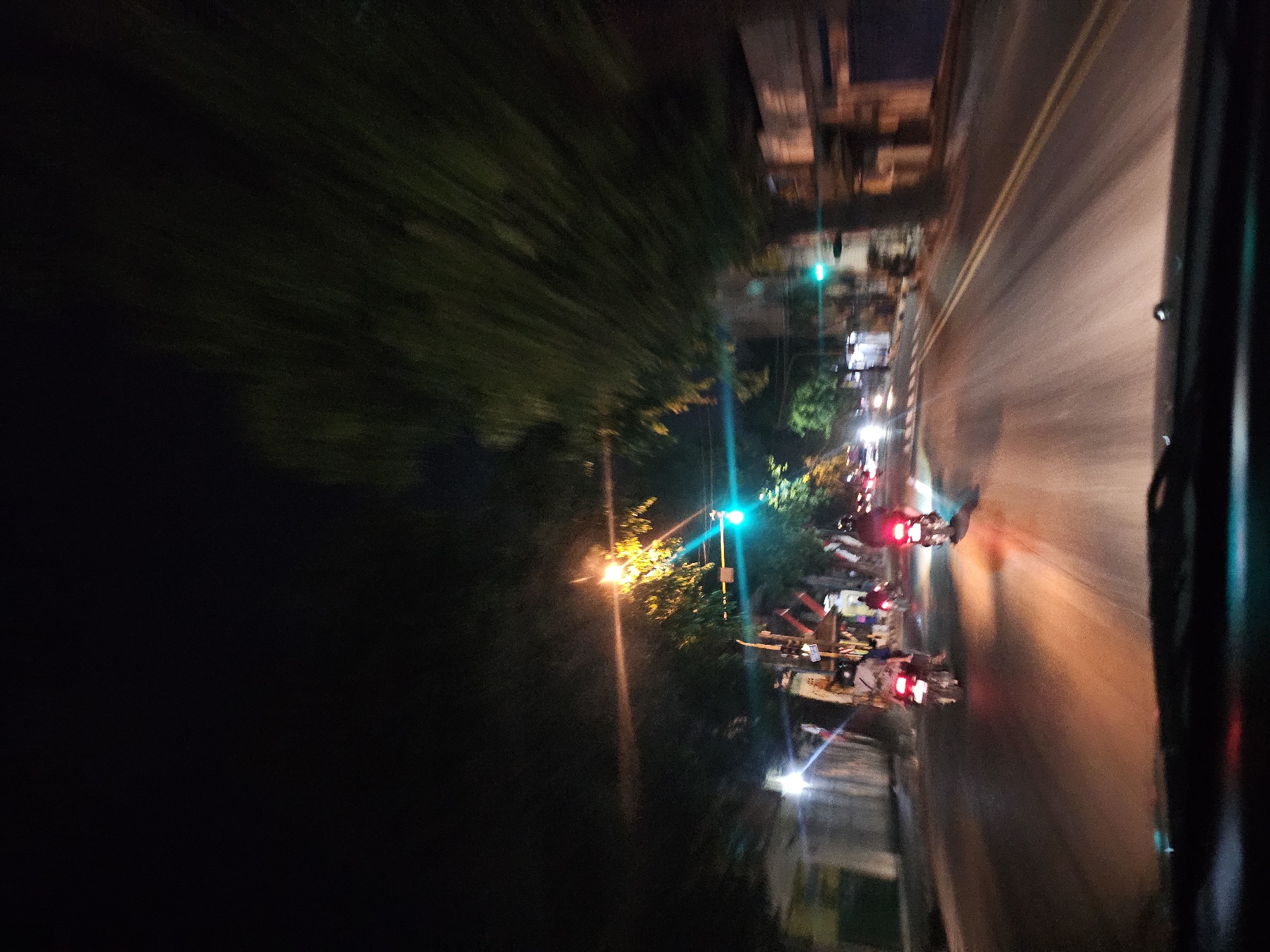 The image depicts a night scene on a city street, likely in Tuban, Indonesia, viewed from inside a vehicle. The road has two lanes, with yellow lines marking the center. Several motorcycles are traveling on the road, their taillights illuminated and casting red and orange reflections on the asphalt. A traffic light is visible, showing a green signal. Trees line the street, their foliage blurred due to motion. Buildings are visible on the right side of the road. The overall impression is one of movement and a typical urban environment at night.