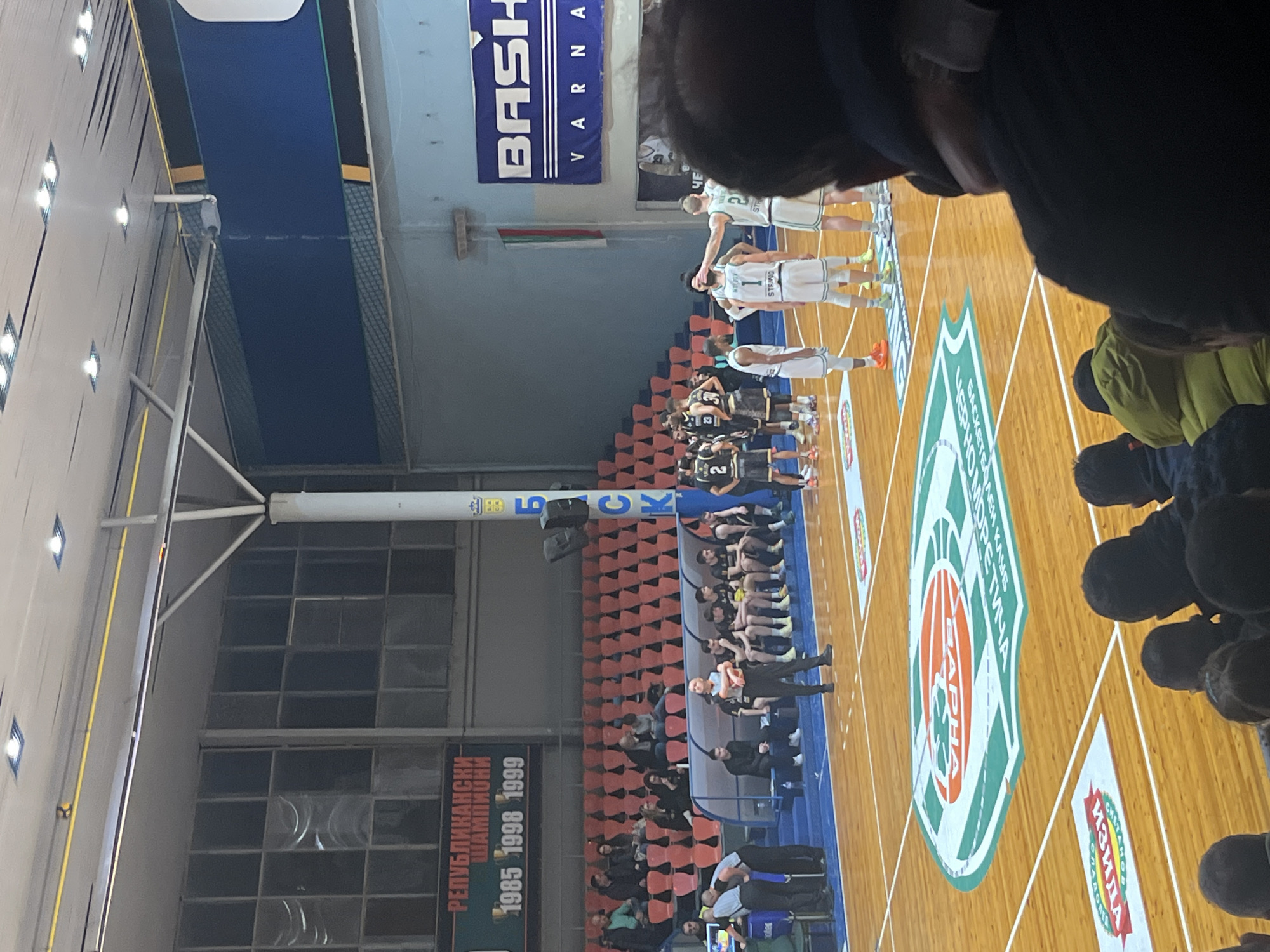 The image shows a basketball game at a sports arena, possibly in Varna, Bulgaria based on the "VARNA" lettering. There are two teams lined up on the court, one in white jerseys and the other in black jerseys. Some players are interacting with each other. Spectators are present in the stands. The court has logos and markings, including text in Bulgarian and logos of sponsors. A banner with "BASH VARNA" is visible, suggesting the team from Varna is playing. The arena has red seating, windows, and structural supports.