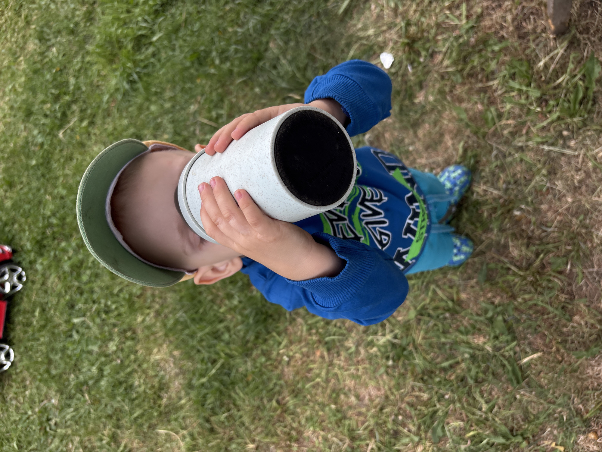 The image shows a child, likely a toddler, standing in grass in Chaikovsky, Russia. The child is wearing a green baseball cap, a blue long-sleeved shirt with green and black designs, and blue and green shoes. The child is holding a cylindrical object to their eye as if it were a telescope or binoculars. The object is light gray with a dark-colored (possibly black) interior visible at the end. A red toy truck is partially visible near the bottom left corner of the image. The environment consists mostly of green grass. The lighting appears to be natural daylight.