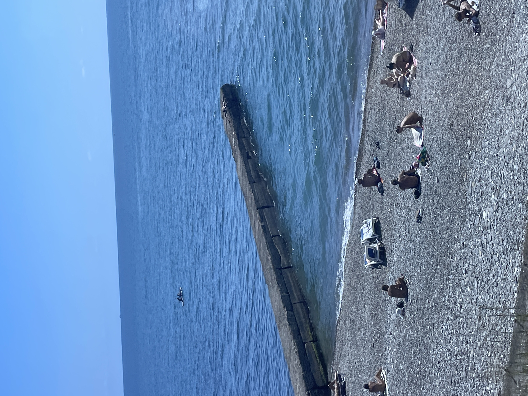 A wide-angle, eye-level shot captures a sunny day at a pebble beach in Sochi, Russia. The calm, blue sea stretches to the horizon under a clear sky. A long, concrete breakwater juts out from the shore, its rough texture contrasting with the smooth water. On the beach, several people are sunbathing on towels and loungers. Some are seated, others are lying down, enjoying the warm weather. Two figures are visible in a small boat in the distance, adding a touch of activity to the serene seascape.