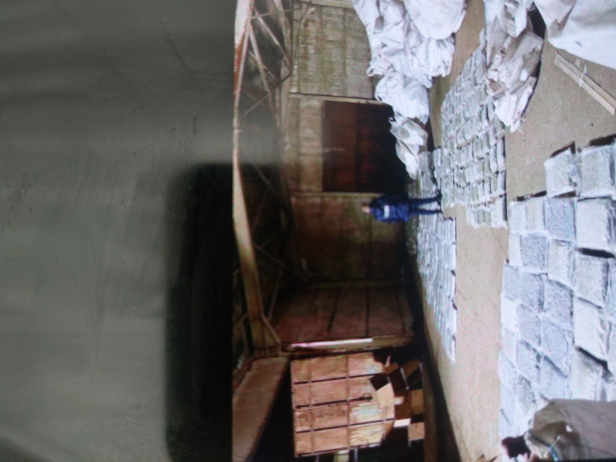 The image appears to be a still from a video or photograph taken inside a warehouse or large storage facility. Located in Tsarevo, Bulgaria. The scene is divided into two main sections:
The left side shows the interior of the building, possibly a storage area, with metal framework and aged walls. There is a stack of wooden crates or boxes in the lower part.
The right side displays a large number of rectangular packages or bundles, arranged in rows. There are also white bags. A man is standing amongst them in a dark blue jacket.