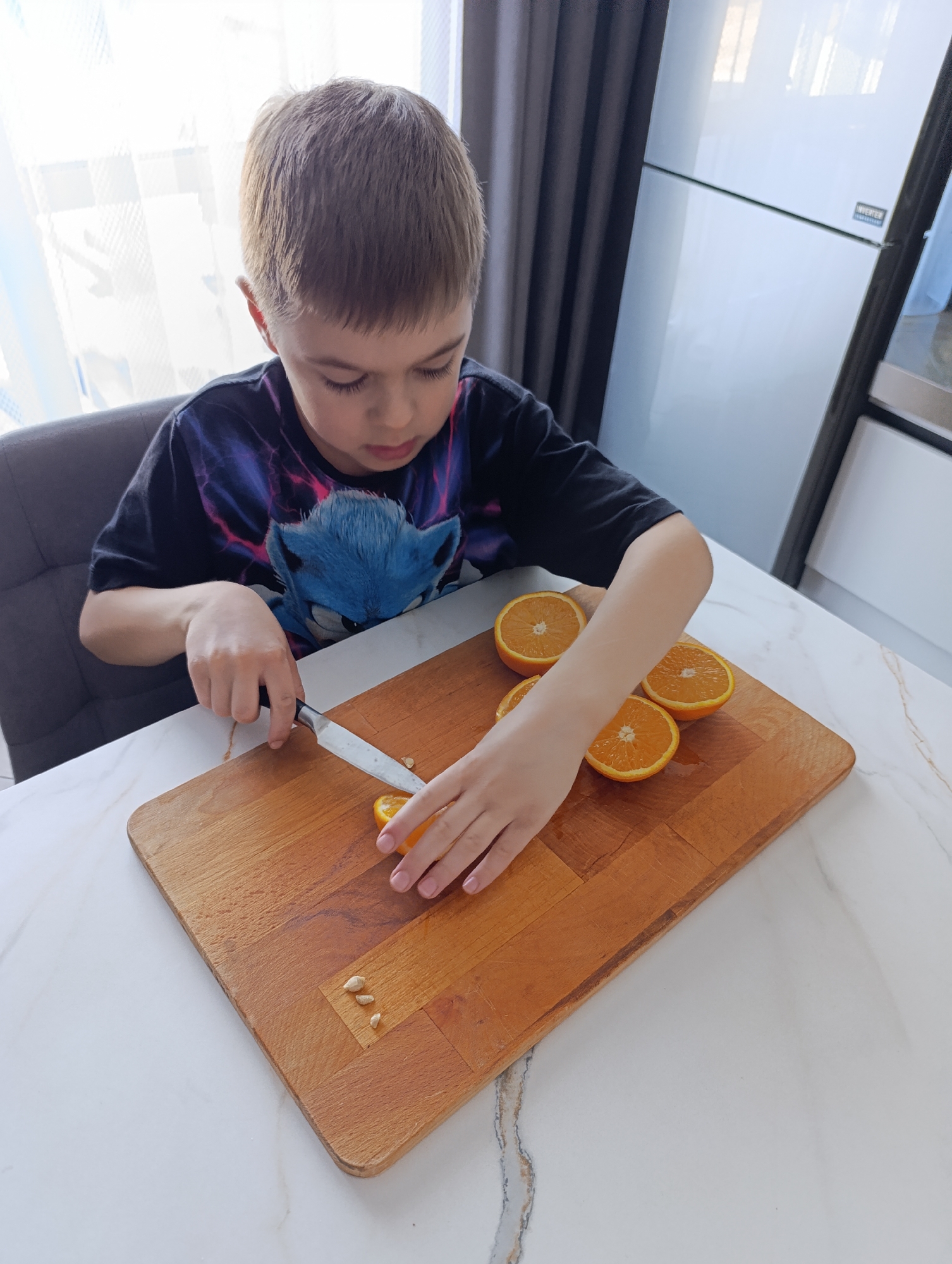 The image captures an indoor scene in Chaikovsky, Russia, likely a kitchen or dining area. A young male child, approximately 6-10 years old, with short light brown hair, is seated at a white marble-patterned countertop. He wears a black short-sleeved t-shirt with a blue graphic, identified as Sonic the Hedgehog. The child is actively engaged in cutting oranges on a rectangular wooden cutting board. His right hand holds a silver-bladed utility knife with a black handle, positioned to slice an orange segment. His left hand stabilizes the orange segment on the board. Four orange halves and one smaller orange segment are present on the cutting board. Three small white orange seeds are visible on the board to the immediate left of the orange segment being cut. In the background, a brightly lit window with white curtains is visible to the left of the child, and a white, two-door refrigerator with an "INVERTER" label is seen to his right. A portion of a gray upholstered chair is visible behind the child.
