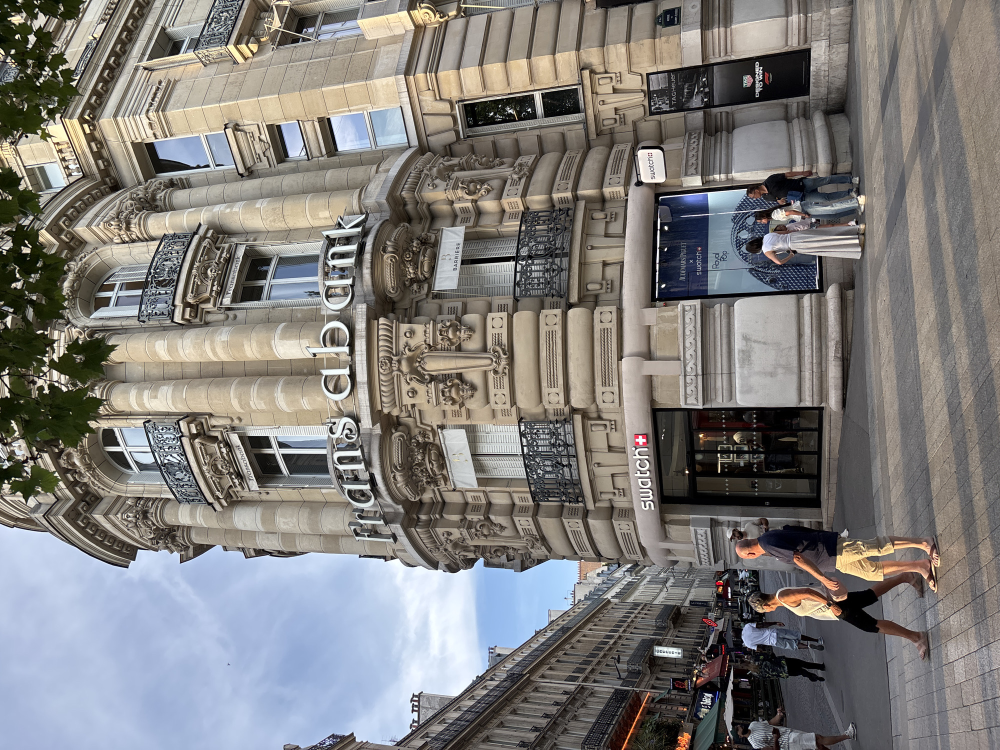 The image shows a street-level view of a corner building in Paris, France, with a bright blue sky and white clouds overhead. The building features ornate architectural details, including columns, balconies with decorative iron railings, and sculpted stonework. Large white letters spelling out "fransabank" are visible on the upper facade. Below, on the ground floor, a Swatch store is prominently displayed with a large window showcasing watches. A few people are walking on the sidewalk and crossing the street in front of the building. The street is lined with other buildings, suggesting an urban environment. The lighting indicates it's late afternoon or early evening, with the sun casting shadows.