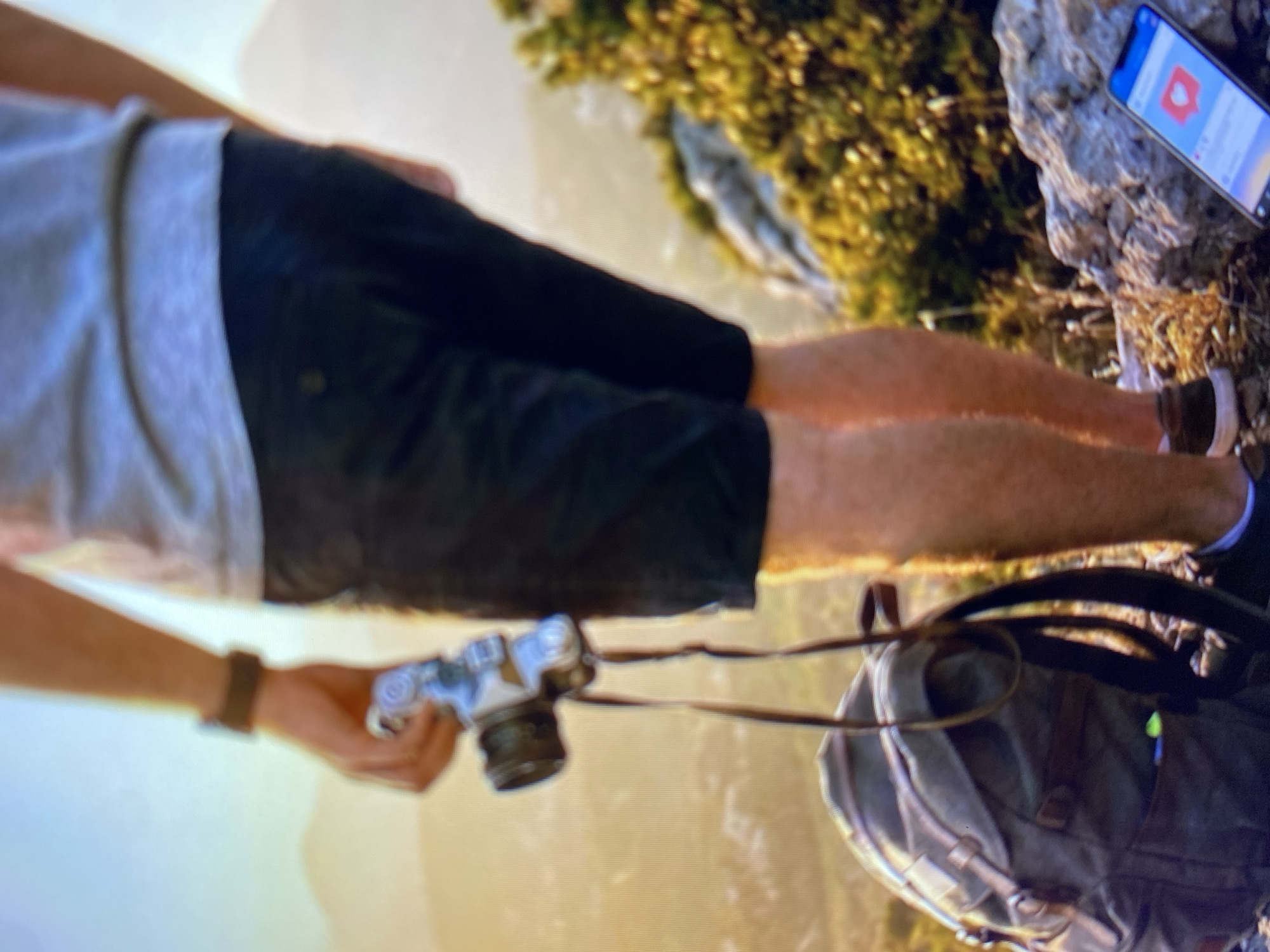 A person, seen from the mid-thigh down, stands on a rocky, overgrown hillside, presumably in Kuta Selatan, Indonesia. The individual is wearing black shorts, a grey t-shirt, and dark sneakers. They are holding a silver digital camera with a black lens and strap. A dark canvas backpack with leather accents rests on the ground to their left. A smartphone, displaying a social media feed with a heart-shaped "like" icon, is positioned on the rocks to the right. The background features hazy, mountainous terrain bathed in warm sunlight.