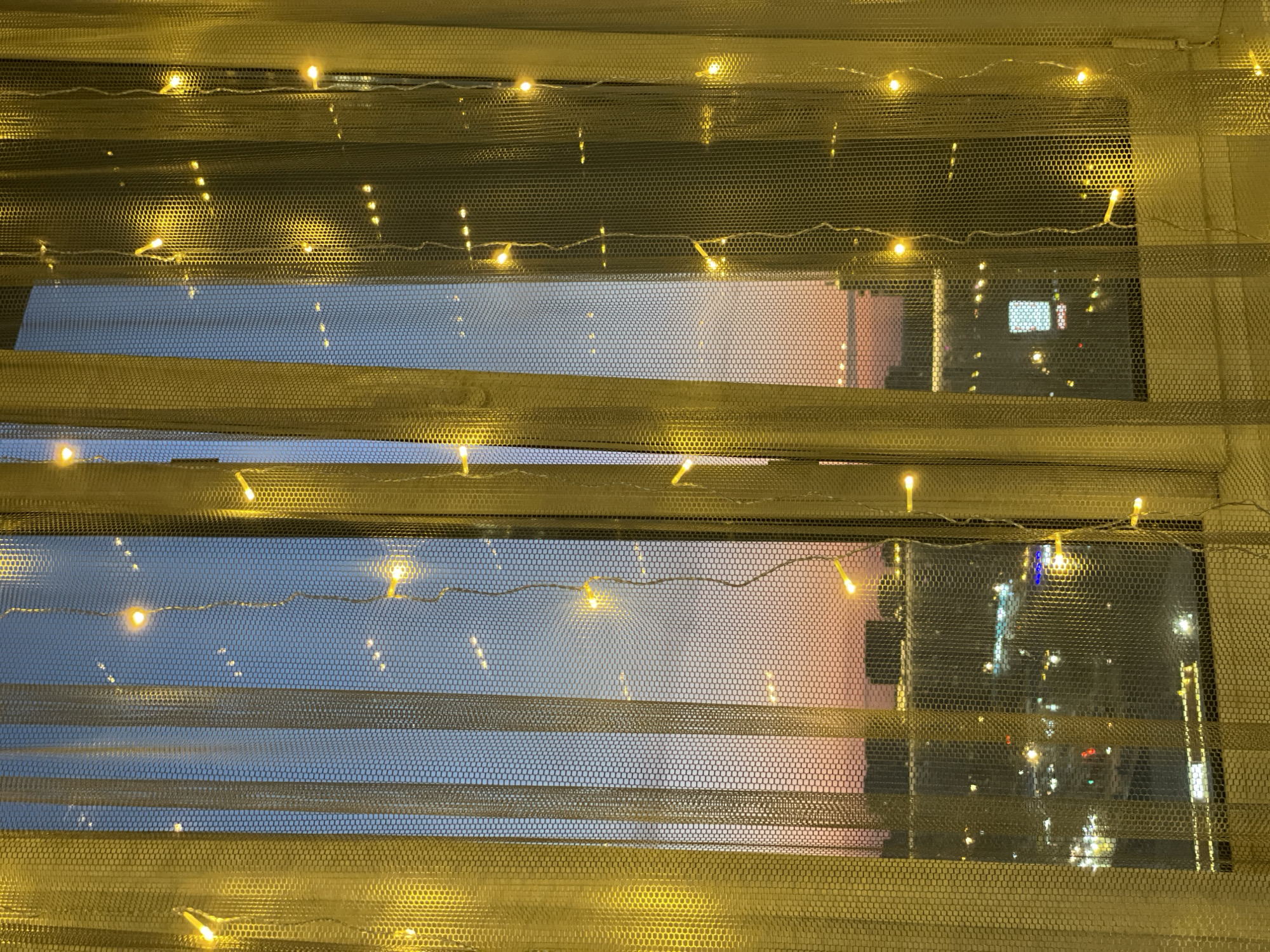 A window is covered by a gold net material decorated with strings of fairy lights, creating a warm, blurred view of the outside. The view reveals the fading light of dusk or dawn in Krasnoyarsk, Russia, with a gradient of blue sky transitioning to a soft orange/pink hue above the horizon. The silhouette of buildings is visible through the netting, and the lights of a distant city are blurred but detectable. The window is divided by horizontal blinds, slightly open, adding layers to the urban landscape.