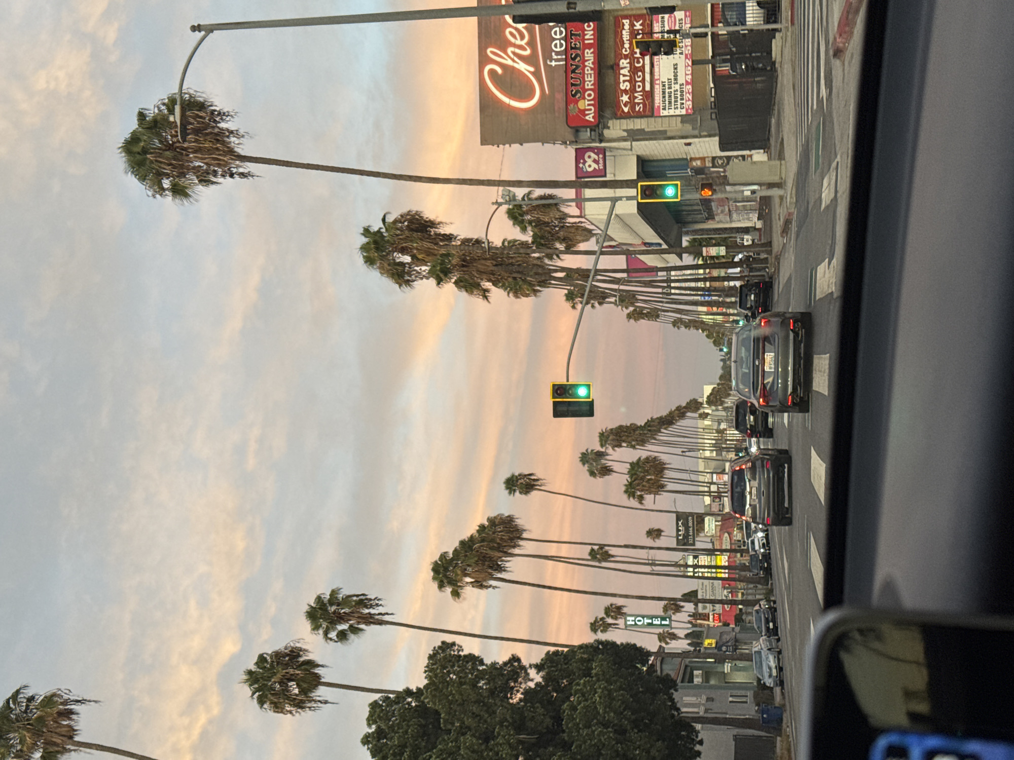 The image captures a street scene in Los Angeles, USA, likely along Sunset Boulevard, characterized by a long row of palm trees lining both sides of the road. The sky displays a soft, pastel gradient of pink, orange, and pale blue, indicating either sunrise or sunset. Multiple cars are driving on the street, with some stopped at a traffic light. A neon sign with the word "Che" is visible on the right side of the road, partially obscured by palm trees and buildings. Signs for "Sunset Auto Repair Inc." and "Star Certified Smog Check" are also present. The perspective is from inside a vehicle, looking forward through the windshield. The overall atmosphere suggests a typical urban environment in Los Angeles during twilight.
