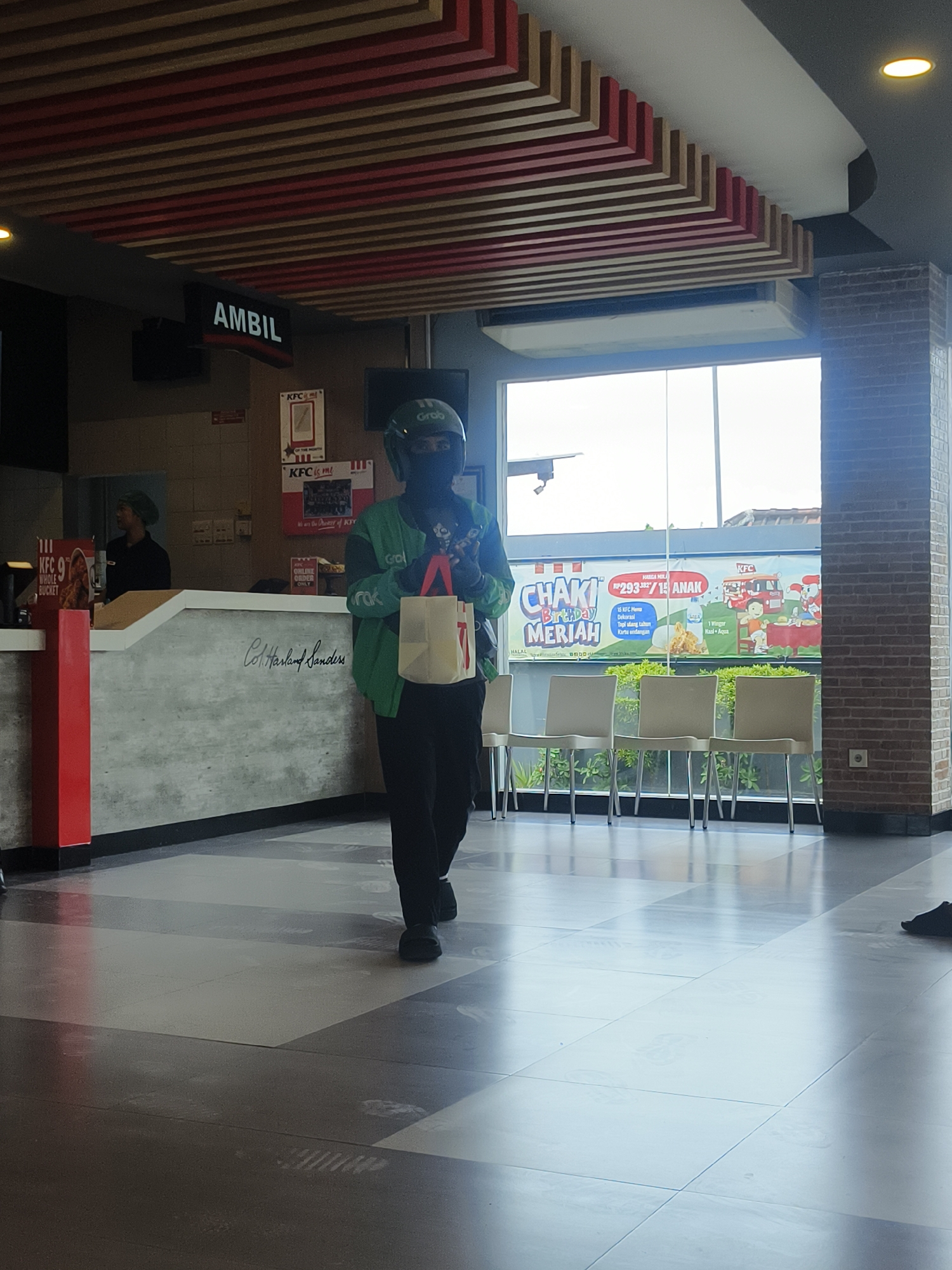 A person wearing a green Grab uniform and a helmet is walking through a KFC restaurant. The person is holding a white paper bag with a red handle. The restaurant has a counter with a sign that says "AMBIL" above it. There are chairs and tables in the background, and a window looking out onto a street. The floor is tiled in a pattern of light and dark gray squares. There is also a KFC logo visible on the wall. The scene appears to be in Denpasar, Indonesia, given the presence of a Grab driver and the Indonesian text on some of the signage.