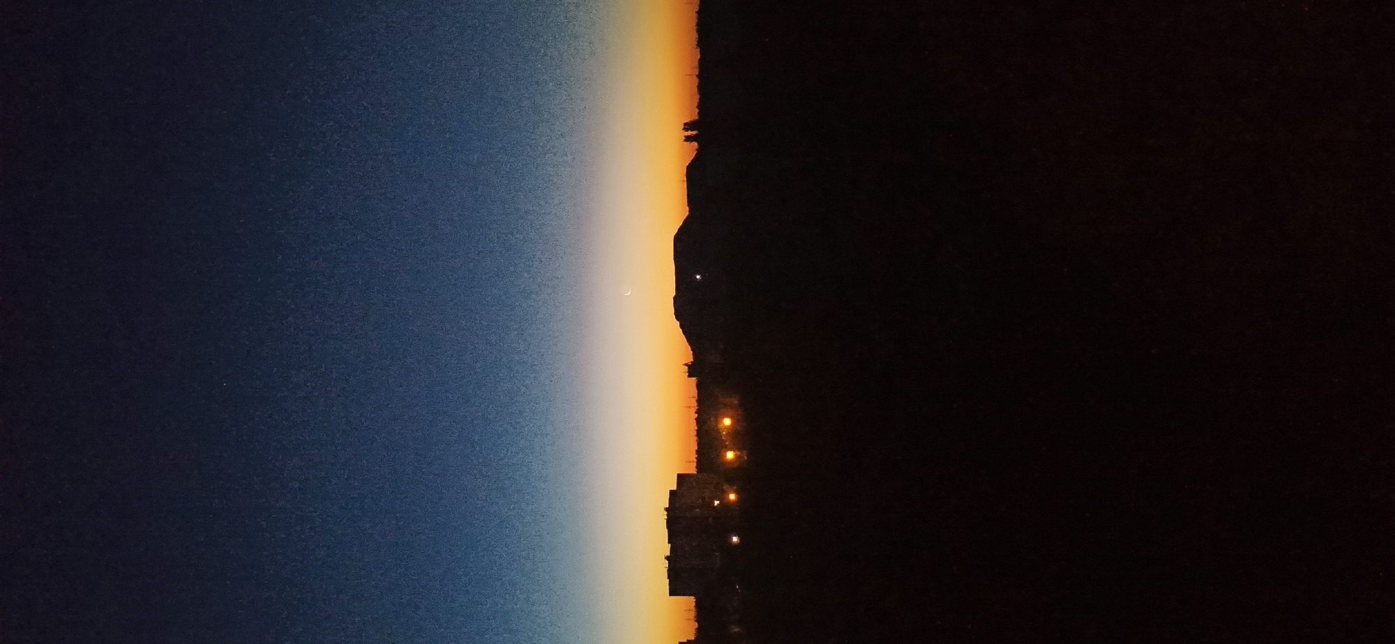 A vertical, low-light, full shot depicts a silhouette of a city skyline during twilight, with a bright crescent moon visible in the sky. The sky transitions from a deep navy blue at the top to a vibrant orange and yellow gradient near the horizon. In the foreground, the silhouette of the ground is entirely black, obscuring any details of the terrain. Silhouetted buildings, some with visible lights, are present in the mid-ground. To the right of the buildings, a prominent, rounded hill or mound is silhouetted against the bright sky. A thin, bright crescent moon is positioned above and slightly to the right of the peak of this hill. The overall impression is one of a calm, transitional moment between day and night in an urban landscape.