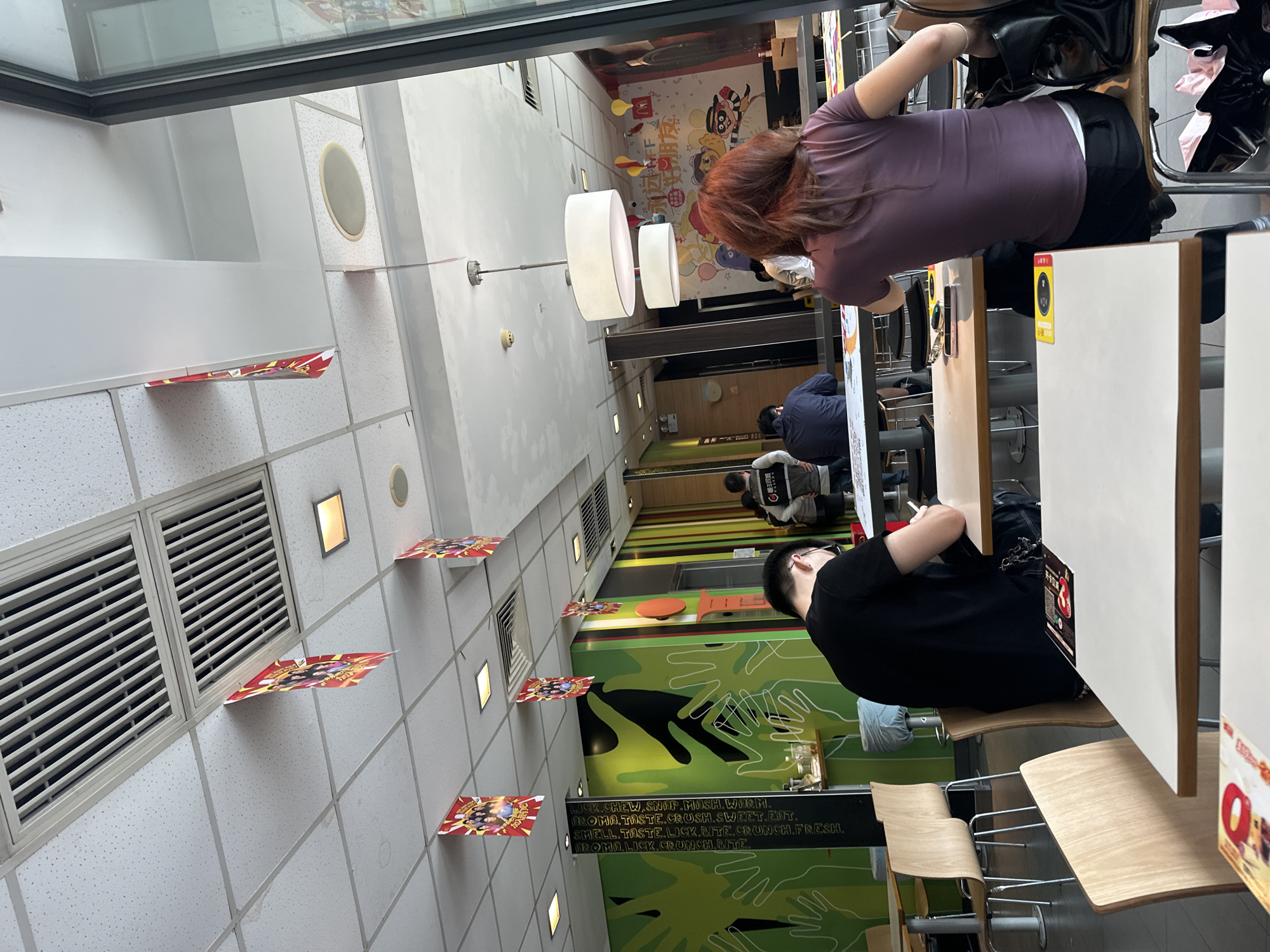 A medium-angle interior shot shows a McDonald's restaurant in Chongqing High-tech Zone, China. The restaurant interior is brightly lit, featuring white-tiled walls adorned with promotional materials. Seated at the tables are several individuals: a woman with auburn hair wearing a purple shirt, and a person in a black shirt and glasses. They appear to be seated and engaged with personal belongings at the table. Other people are visible further in the background. The restaurant is decorated with whimsical, hand-drawn-style graphics on the walls, and is equipped with modern lighting fixtures. The scene suggests a casual dining environment.