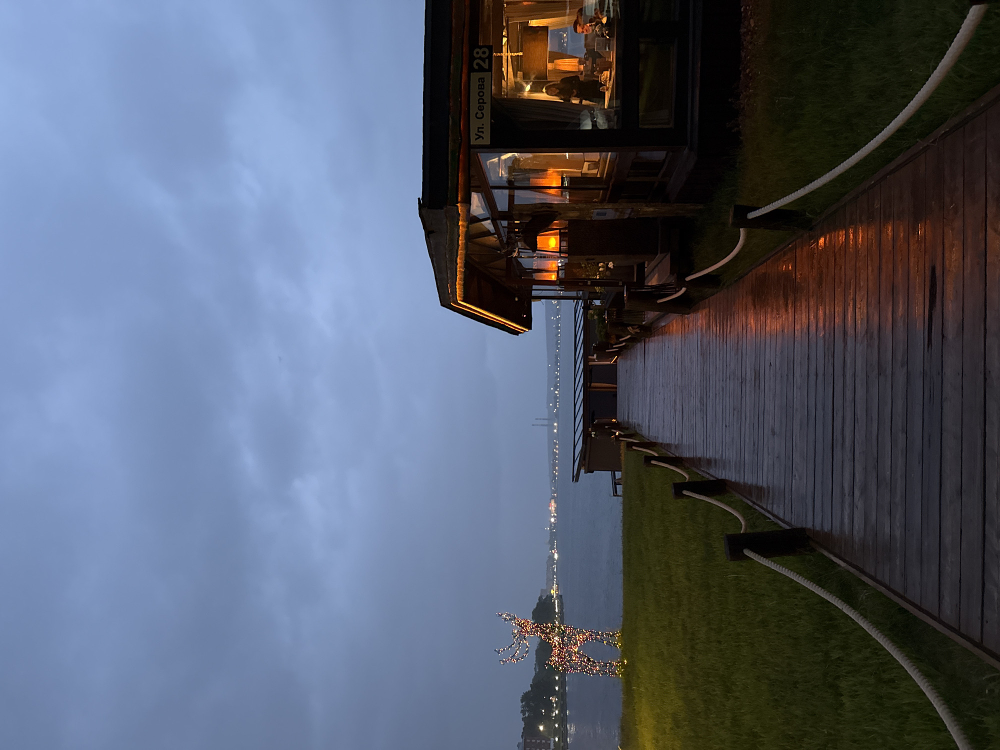 The image depicts an outdoor twilight scene, likely after rainfall, centered on a wet wooden boardwalk leading towards a body of water. The location is identified as Serov Street 28 (Ул. Серова 28) in Votkinsk, Russia, corresponding to a waterfront establishment.
In the immediate foreground, a dark, wet wooden boardwalk extends from the bottom of the frame towards the midground. It is flanked on both sides by rope railings supported by dark wooden posts. The wet surface of the boardwalk reflects warm, orange light originating from the building on the right.
To the right of the boardwalk is a modern single-story building, likely a restaurant or cafe, with large glass windows. The building features dark framing and roofline, accented by orange LED strip lighting along its eaves. Interior lighting within the building is warm and bright, illuminating several individuals visible through the windows; at least one person appears to be standing, and another is seated at a table.
To the left of the boardwalk, a patch of vibrant green grass extends. On this grass stands a large, illuminated decorative sculpture of a reindeer, composed of multi-colored lights (red, green, blue, yellow, etc.).
Beyond the boardwalk and the immediate shoreline, a wide body of water, likely the Votkinsk Reservoir, stretches into the distance. The water surface appears calm under the overcast sky. Across the water, the distant shoreline of Votkinsk is visible, punctuated by numerous small, twinkling artificial lights. Industrial structures, including what appear to be smokestacks or towers, are faintly discernible on the far horizon against the dim light.
The sky is overcast and uniformly grey-blue, indicating twilight or heavily cloudy conditions, with no direct sunlight visible. The overall ambiance is muted and cool, with localized pockets of warm artificial light.