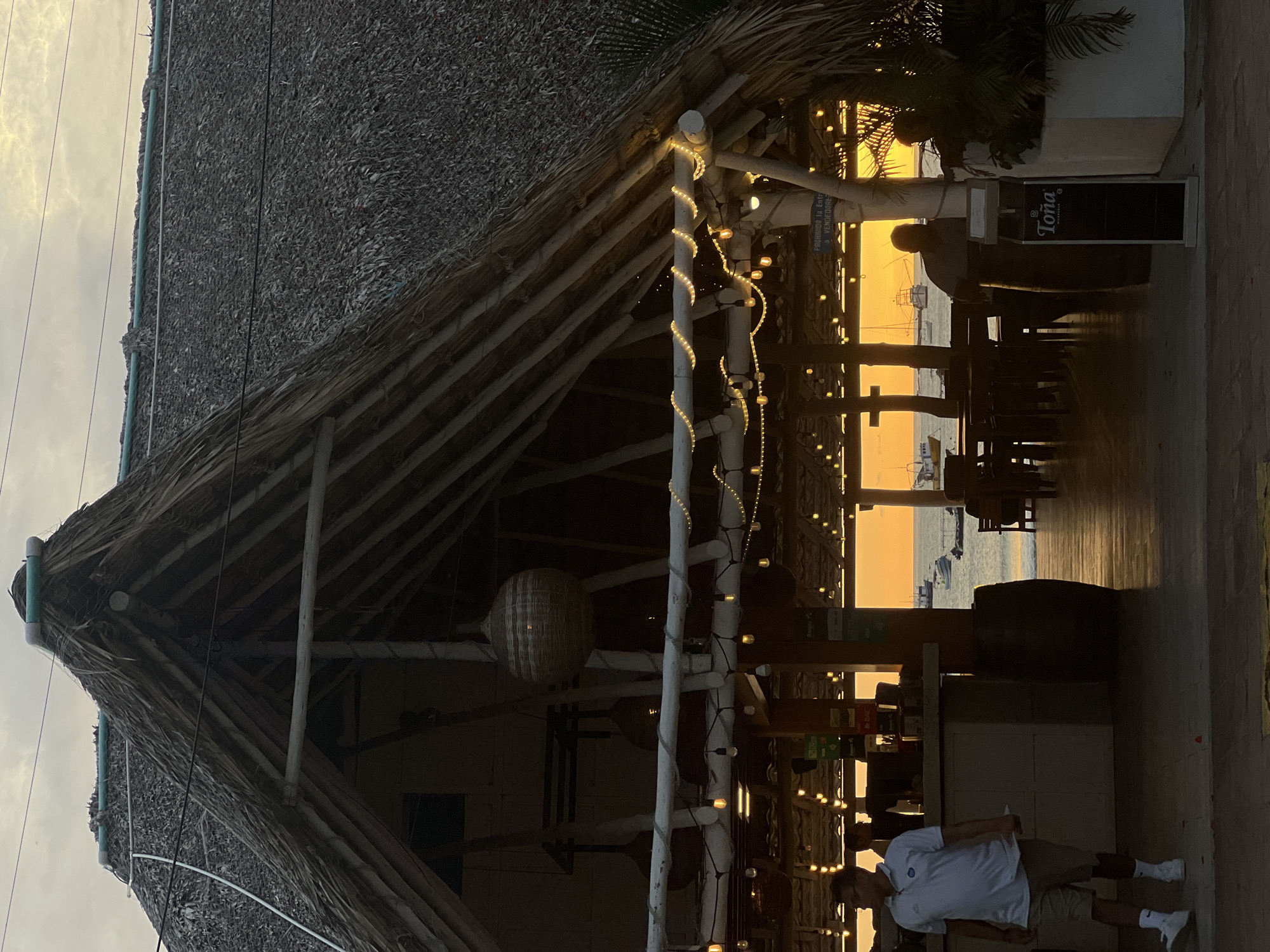 The image depicts a beachfront establishment in San Juan del Sur, Nicaragua, at sunset. The foreground is dominated by a thatched-roof structure with an open-air seating area. A man in a white polo shirt and tan shorts is walking past a bar area. The bar has various payment signs displayed. Behind the bar, through the open structure, a view of the ocean is visible with several boats anchored. The setting sun casts a warm orange glow over the scene. The building features decorative string lights along its roofline. In the background, more tables and chairs are set up, and people are dimly visible inside the establishment. The overall atmosphere suggests a relaxed, tropical evening.