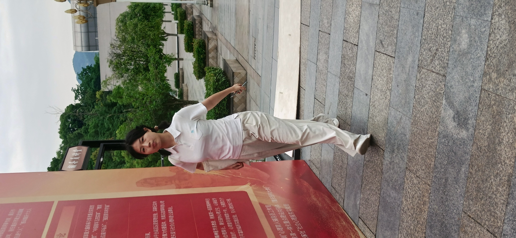 A woman with black hair in two pigtails is standing in an outdoor paved area. She is wearing a white polo shirt and light beige pants. Her right arm is extended, holding a small dark object. She is looking at the camera with a neutral expression.
Behind her is a large informational sign with Chinese characters and some red tones. To the left, some trees and greenery are visible. In the background, part of a building is visible, as well as what appears to be a mountain or hill in the distance. The location is identified by the text on the sign as the city of Chongqing High-tech Zone, China.