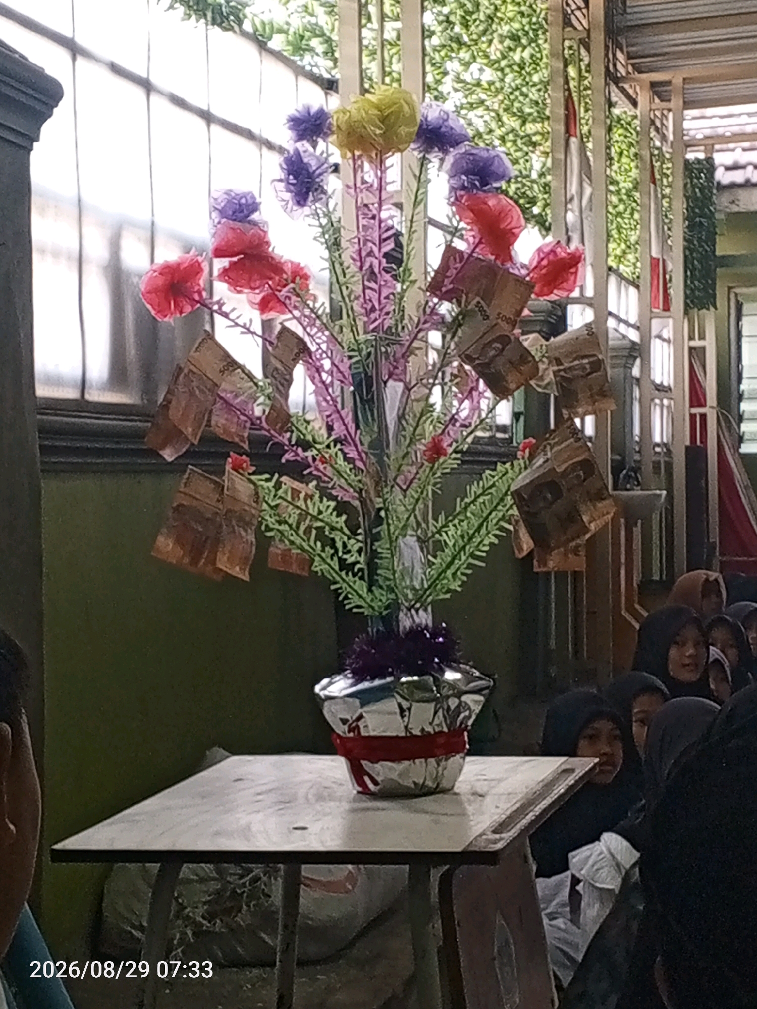 A decorative arrangement of artificial flowers and money hangs on display in Tanjungan, Indonesia. The centerpiece features a bouquet with a vibrant assortment of purple, pink, and yellow artificial blossoms. Several folded banknotes, appearing to be Indonesian Rupiah, are attached to the branches of the arrangement. The bouquet is housed in a silver, crinkled vase adorned with a red ribbon. This arrangement sits on a light-colored tabletop, which is supported by a wooden structure. In the background, a group of children, mostly wearing hijabs, are visible, observing the display. The environment suggests an indoor or covered outdoor setting, with natural light filtering in from the left. The overall scene appears to be part of a cultural or celebratory event.