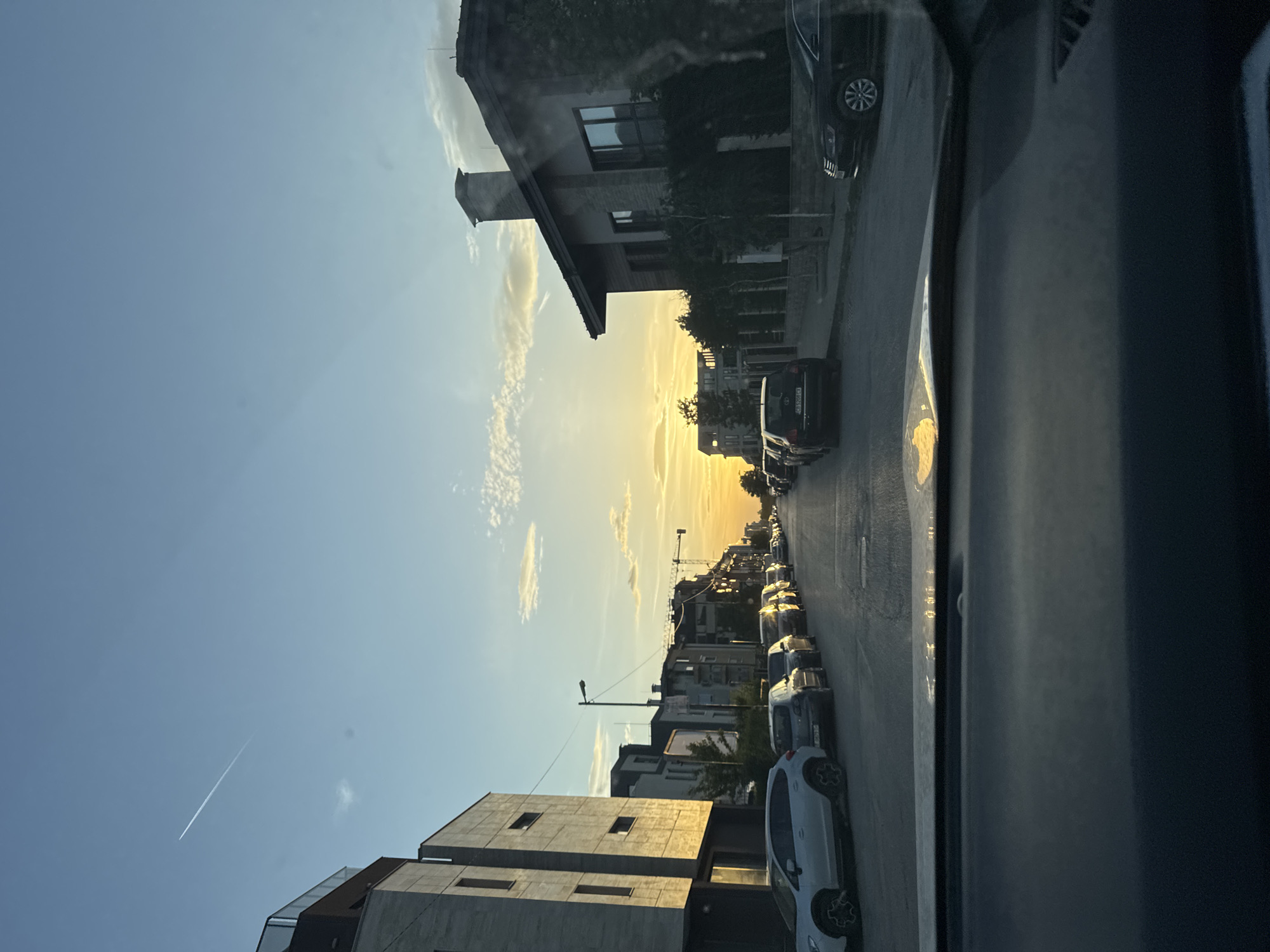 Captured from inside a car, the image shows a street scene at dusk in Sofia, Bulgaria. The viewpoint is from the driver's seat, looking towards the setting sun which casts a golden light down the street. Cars line both sides of the road. Buildings are visible on either side, a mix of residential and commercial structures. In the sky, there is a contrail from an airplane.