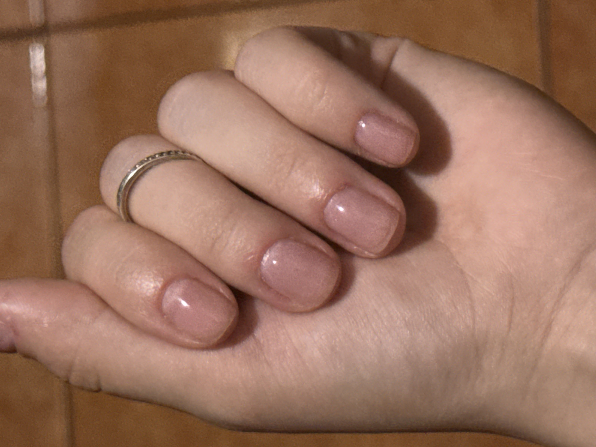 A close-up, high-angle shot shows a person's left hand, which is resting on a textured, wood-like surface, likely a floor. The hand is adorned with a thin, silver-toned ring on the ring finger and has neatly manicured fingernails painted with a sheer, pale pink polish. The lighting suggests an indoor environment. There is no indication of the image's location within Lobacheve, Ukraine.
