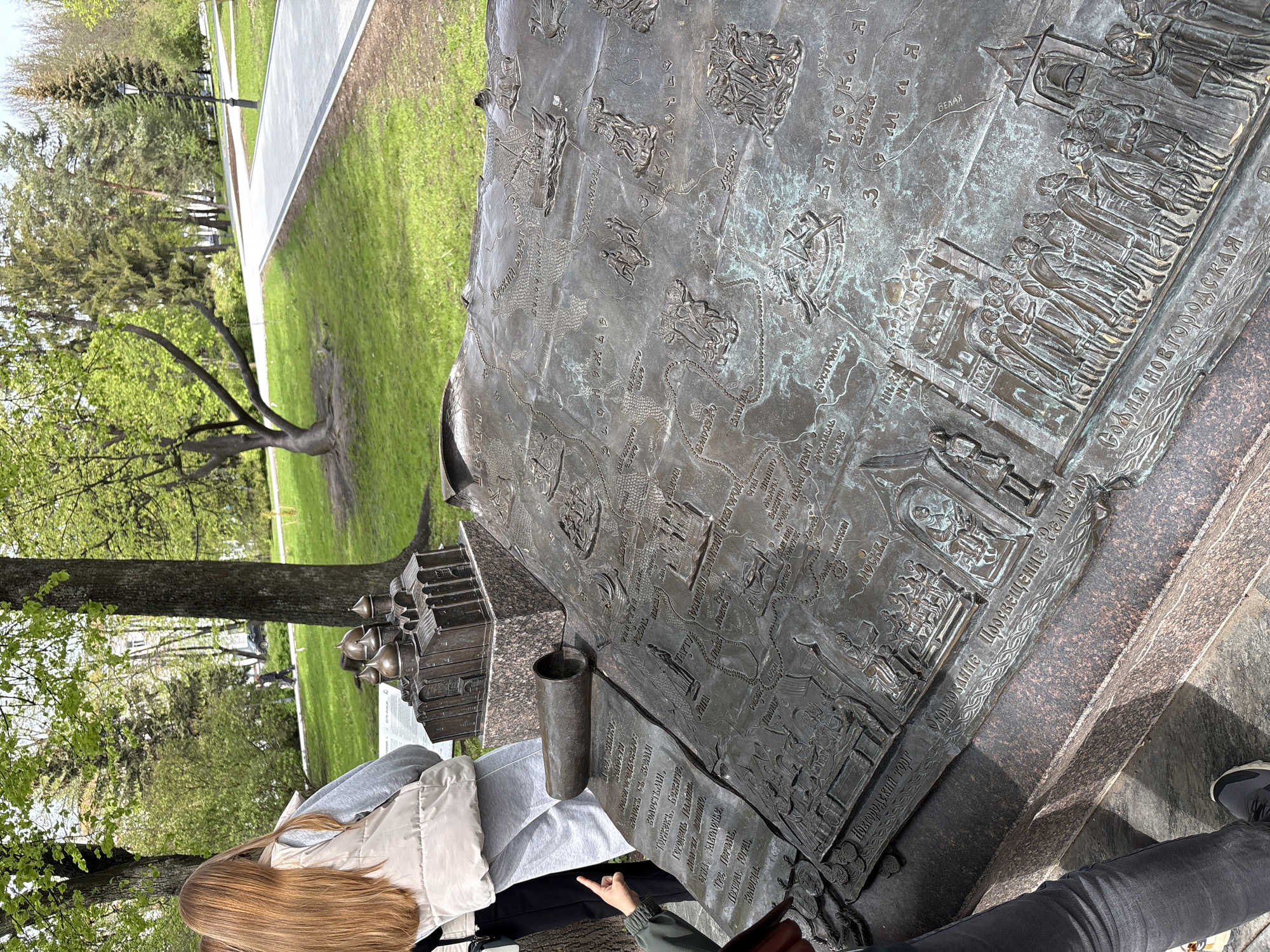 Here is a detailed description of the image:
The image shows a bronze monument or sculpture in a park-like setting, likely in Veliky Novgorod, Russia, given the Cyrillic text on the sculpture. The main subject of the image is the textured bronze surface, which features a map-like depiction and bas-reliefs of various scenes and figures. The bronze artwork is set on a stone base and appears to be an interactive display, possibly an informational piece about the history or notable places of Veliky Novgorod.
A woman with long, reddish-blonde hair, wearing a cream-colored vest over a darker top, is seen pointing at a section of the bronze sculpture. There are other people partially visible, including someone wearing dark jeans and sneakers, suggesting visitors are interacting with the monument. The bronze artwork has names such as "Moscow", "Yaroslavl", "Rostov", "Sofia Novgorodskaya", and “Novgorodsky Torg (Novgorod Trade)” inscribed on its surface.
The surrounding environment shows a park-like setting with green grass, trees with fresh spring leaves, and a pathway in the background. The lighting suggests it is daytime. The scene appears to be an outdoor public area in Veliky Novgorod, offering visitors a historical or cultural point of interest within a green space.