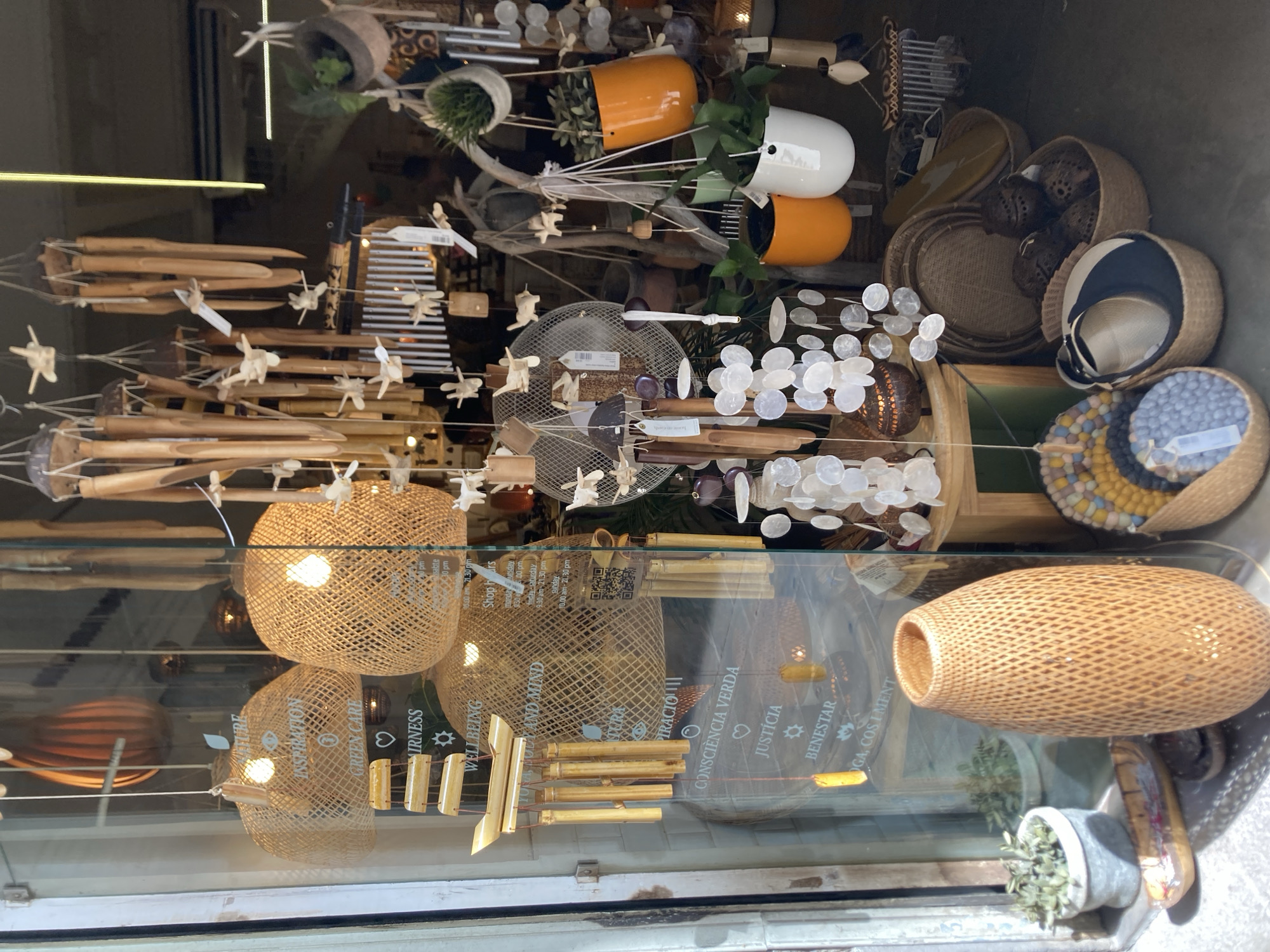 The image displays the interior of a shop, seen through a glass window, likely located in Barcelona, Spain. The shop appears to be a boutique selling artisanal and decorative items, with a strong emphasis on natural materials like bamboo and woven fibers.
Dominating the foreground and midground are several hanging decorative elements. These include numerous wind chimes made from bamboo tubes of varying lengths, some with small carved wooden bird figures attached. There are also strands of round, white shell-like discs hanging, creating a shimmering effect. Two large, dome-shaped lamps made of woven natural fibers hang prominently, each with a visible light bulb inside.
On the left side of the window, text is visible on the glass, appearing to be a list of principles or values such as "NATURE," "INSPIRATION," "GREEN CARE," "WELLBEING," "CONSCIÊNCIA VERDA" (Green Conscience), "JUSTIÇA" (Justice), "BENESTAR" (Wellbeing), and "YOGA, COS I MENT" (Yoga, Body and Mind).
Within the shop's interior, various objects are arranged. A tall, intricately woven basket-like vase stands on the left. In the mid-right, several hanging planters containing green plants are visible, with orange and white ceramic pots. Towards the back, various woven baskets are on the floor, some containing dark, spherical decorative items. A round, colorful mat-like object is also visible in the lower left.
The lighting suggests it is daytime, with light illuminating the shop's interior. Reflections on the glass indicate an outdoor environment beyond the window. The overall impression is of a serene and bohemian retail space.