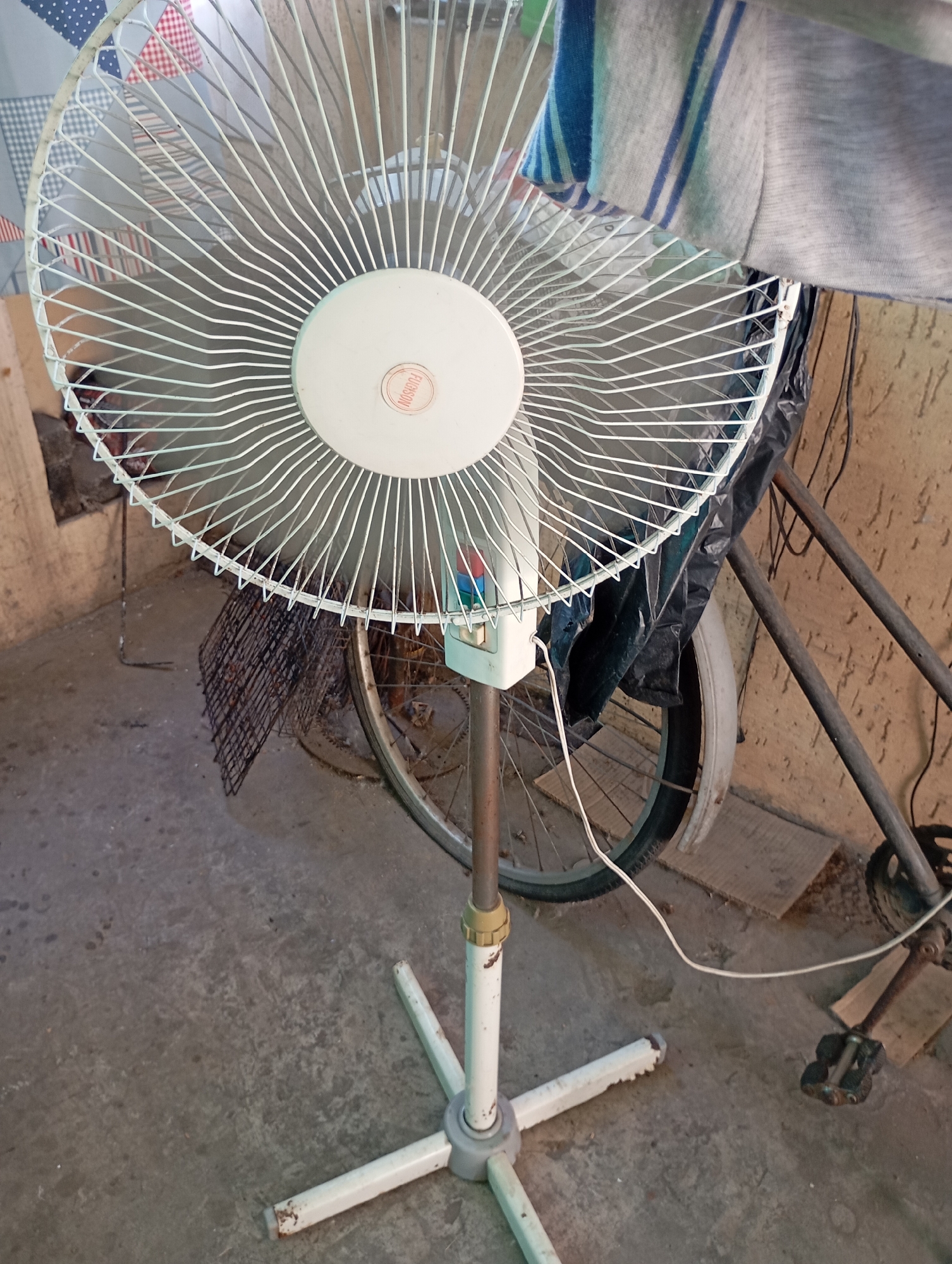 This is a medium-shot, eye-level, indoor photograph of a white standing fan positioned in front of a bicycle and a wall. The fan is white and has a circular grill with a circular logo in the center. The fan is on a white, star-shaped base. The bicycle has a black tire and a silver fender. The wall in the background appears to be concrete or stucco and is a light brown color. There is also some dark plastic sheeting behind the fan. A white power cord is visible running from the fan. The floor is concrete. There are no people in the image. The scene is consistent with a storage area or garage in Horishni Plavni, Ukraine.