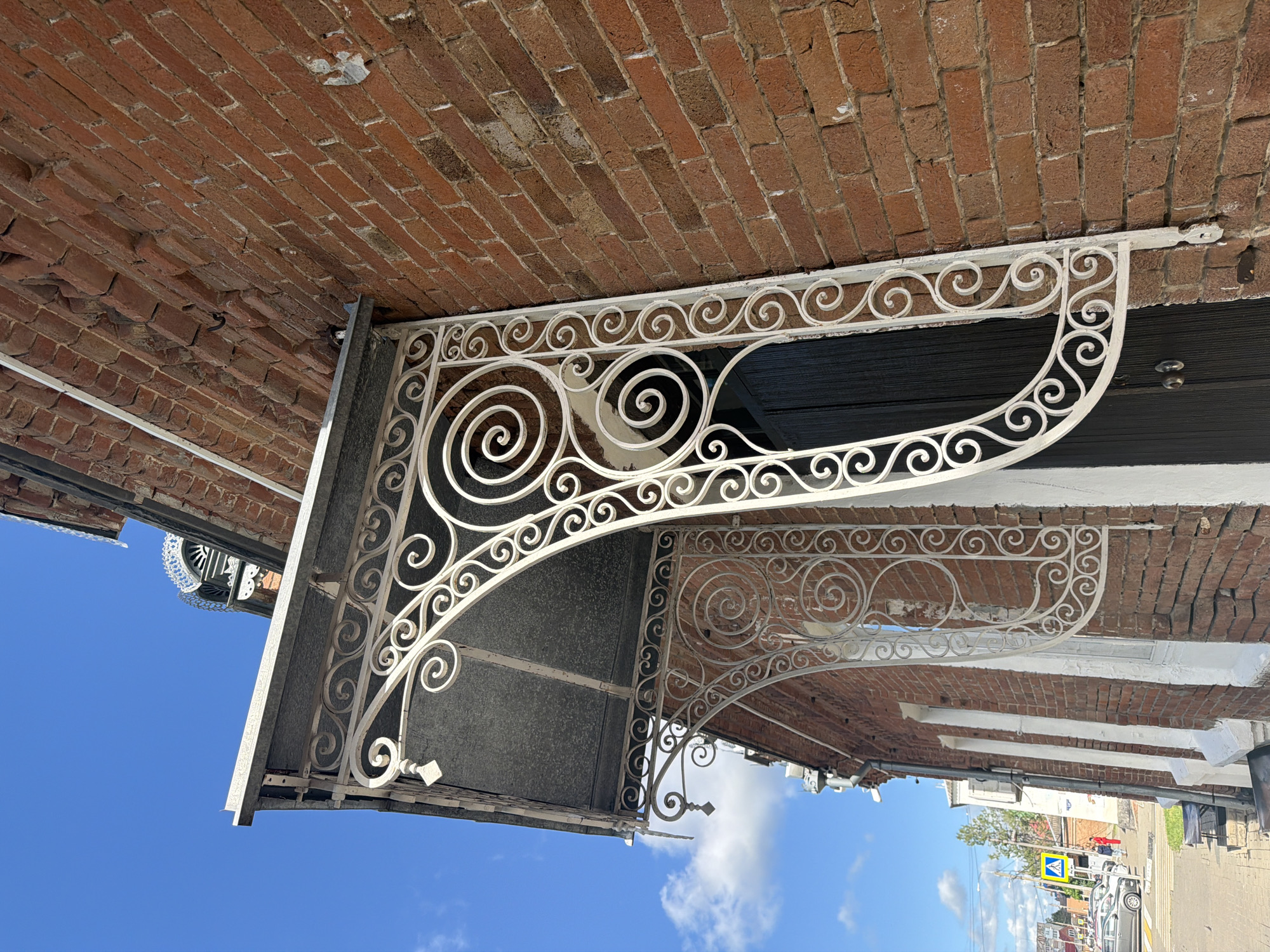 The image shows the exterior of a brick building in Leningradskaya, Russia. It features a white, ornamental iron awning with spiral designs. The building has a dark-colored door visible under the awning. The sky is blue with some clouds. In the background, other buildings, trees, vehicles, and road markings are visible on a street. The image is taken from a low angle, tilting upwards.