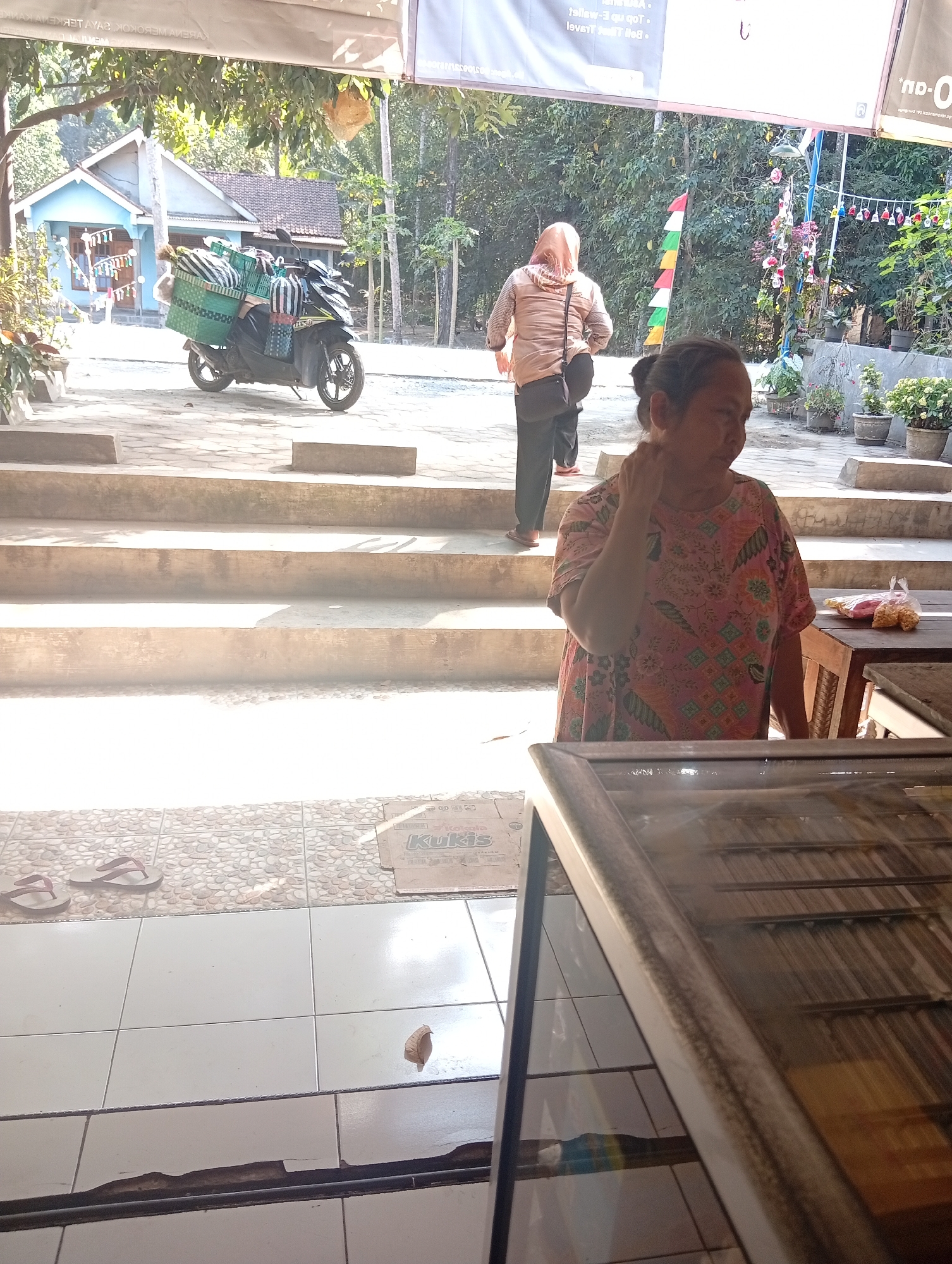 In Plempukankembaran, Indonesia, a woman in a colorful patterned dress is seen standing near a wooden table. She is touching her neck with her right hand. Behind her, another woman wearing a light brown headscarf and a patterned shirt walks away, carrying a dark bag. In the background, a light blue house is visible, with a motorbike parked in front of it. The motorbike is loaded with green baskets and striped bags. The scene appears to be outdoors, with trees and foliage surrounding the area. A set of concrete stairs leads up to the area where the women are standing. Several potted plants are also visible. The ground is a mix of tiled and stone surfaces.