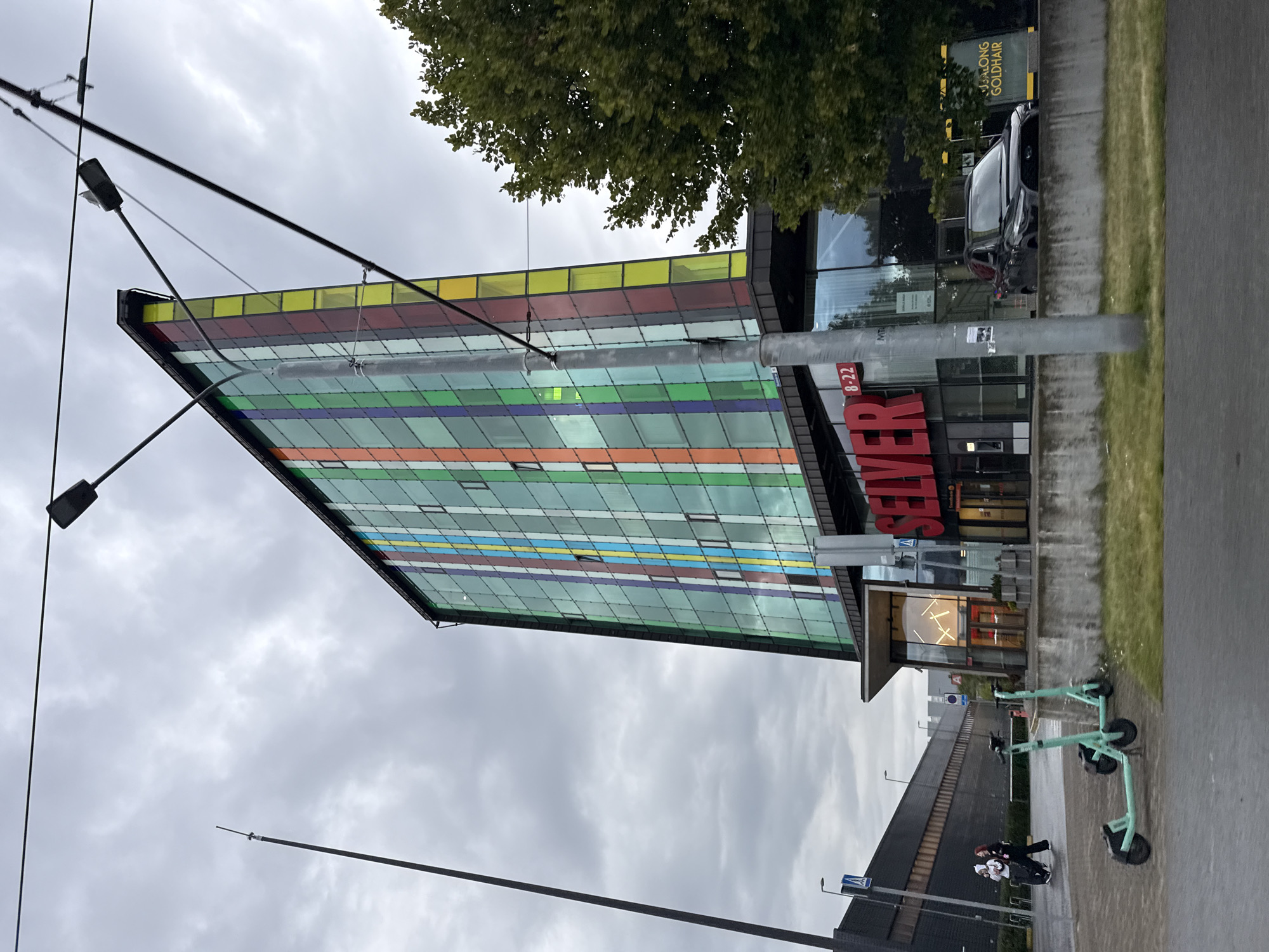 The image depicts a building with a vibrant, multi-colored facade, featuring vertical stripes of red, orange, yellow, green, blue, and purple glass. The building is a modern, multi-story structure with visible windows. In front of the building, there is a concrete barrier and some greenery. Two light-green electric scooters are parked on a paved surface in the foreground. To the left of the building, two people, one wearing a white hooded garment and the other in dark clothing, are walking. Further left, a dark, angled structure with a tall, thin pole rises into the overcast sky. A black car is visible parked near the entrance of the building. A street sign reading "SEVER 8-22" is prominently displayed on the building. The scene suggests an urban environment, likely in Tallinn, Estonia, given the architectural style and the presence of electric scooters, which are common in European cities. The overall atmosphere is somewhat muted due to the cloudy sky.