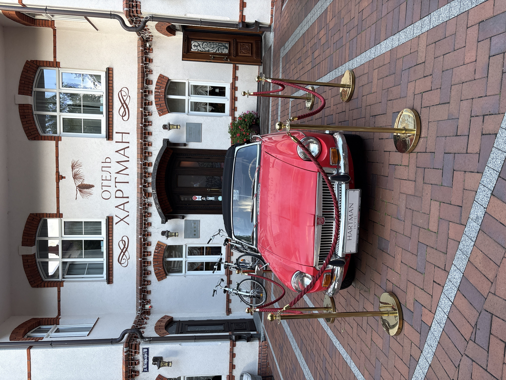In Svetlogorsk, Russia, a bright red convertible car, possibly a vintage MG MGB, is parked on a brick paver driveway in front of a white building with decorative brickwork around its arched windows and doors. The building features a sign that reads "ОТЕЛЬ ХАРТМАН" (Hotel Hartman) in large, stylized letters, with a silhouette of a bird or pine cone above it. Two bicycles are leaning against the building to the left of the entrance. The car is cordoned off by gold stanchions with red ropes, suggesting it is a display or is being protected. The overall scene depicts a charming and well-maintained exterior of a hotel or establishment.