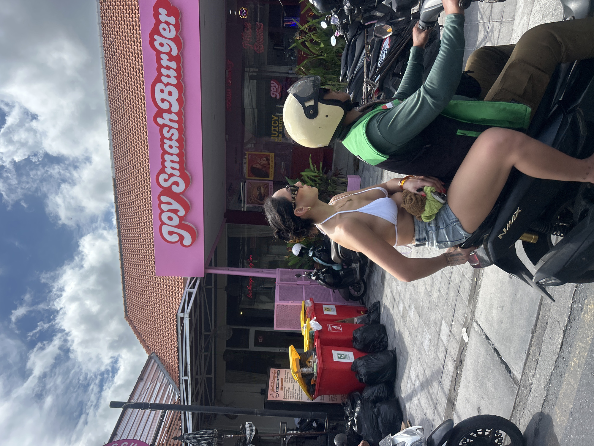 A medium shot captures two individuals riding a scooter on a street in North Kuta, Indonesia. The person in the back, a woman wearing a white bikini top and denim shorts, is looking to the left and holding a small, brown, stuffed animal. The person in the front, wearing a green vest over a dark long-sleeved shirt and a yellow helmet, is driving the scooter. Behind them, a pink building with the text "Joy Smash Burger" is visible, featuring a tiled roof. To the left of the building, several red trash bins with yellow lids are present, alongside black trash bags on the ground. Other scooters are parked in the background. The sky is partly cloudy.