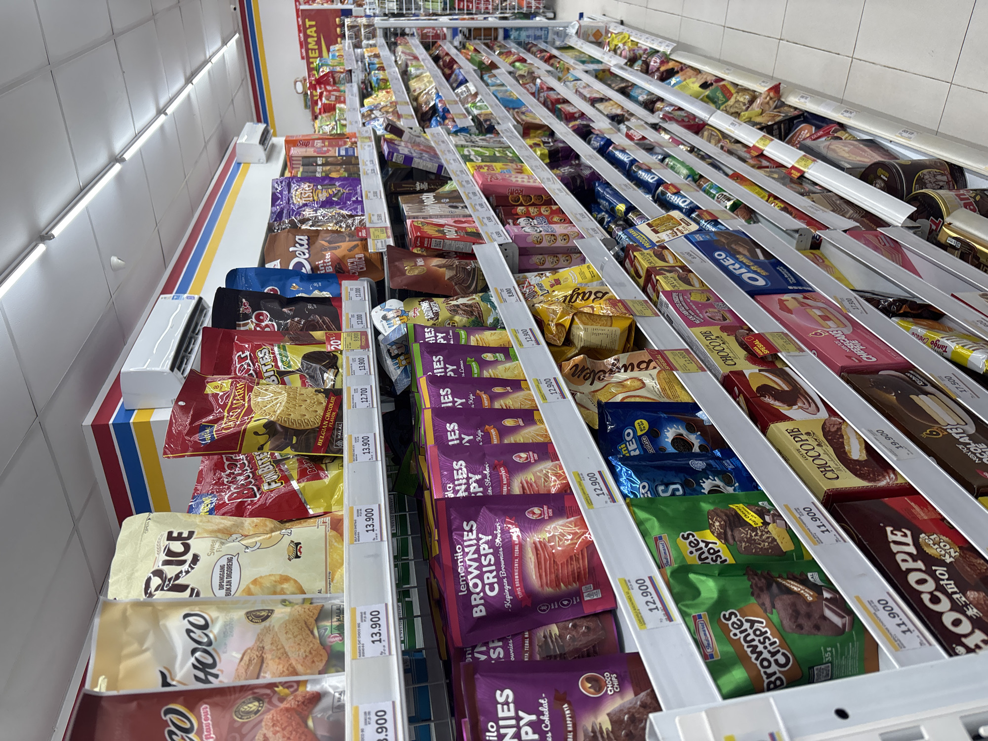 The image shows shelves filled with various snack products in a retail store, likely an Indomaret convenience store, in Cirebon, Indonesia. The shelves are stocked with branded items such as "Hoco," "Rice," "Butter," "Deka," "Niko," "Oreo," and "Chocopie." The products are arranged in colorful packaging, with prices visible on some of the shelf labels. The store appears to be well-lit, with fluorescent lights on the ceiling. The background shows more shelves stocked with snacks and a section with larger promotional items, including a red banner with "HEMAT" written on it, indicating a sale or discount. The floor is tiled with light-colored tiles. There are no people visible in the image.