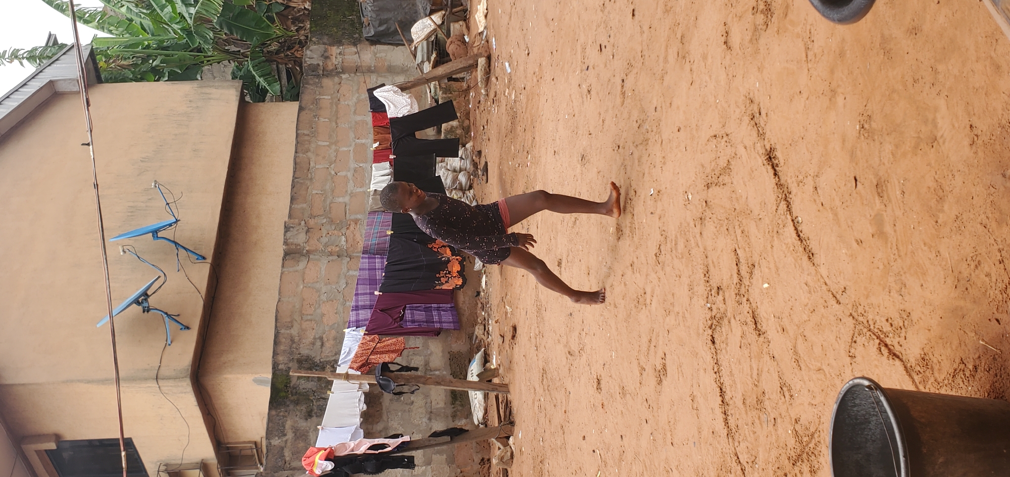 The image captures a scene in Onitsha, Nigeria. A young person stands barefoot on a dirt ground. To the left, clothes hang on a makeshift wooden clothesline. A building with two blue satellite dishes attached to the wall is also visible. Lush green banana trees are seen in the background. A metal bucket sits in the foreground.