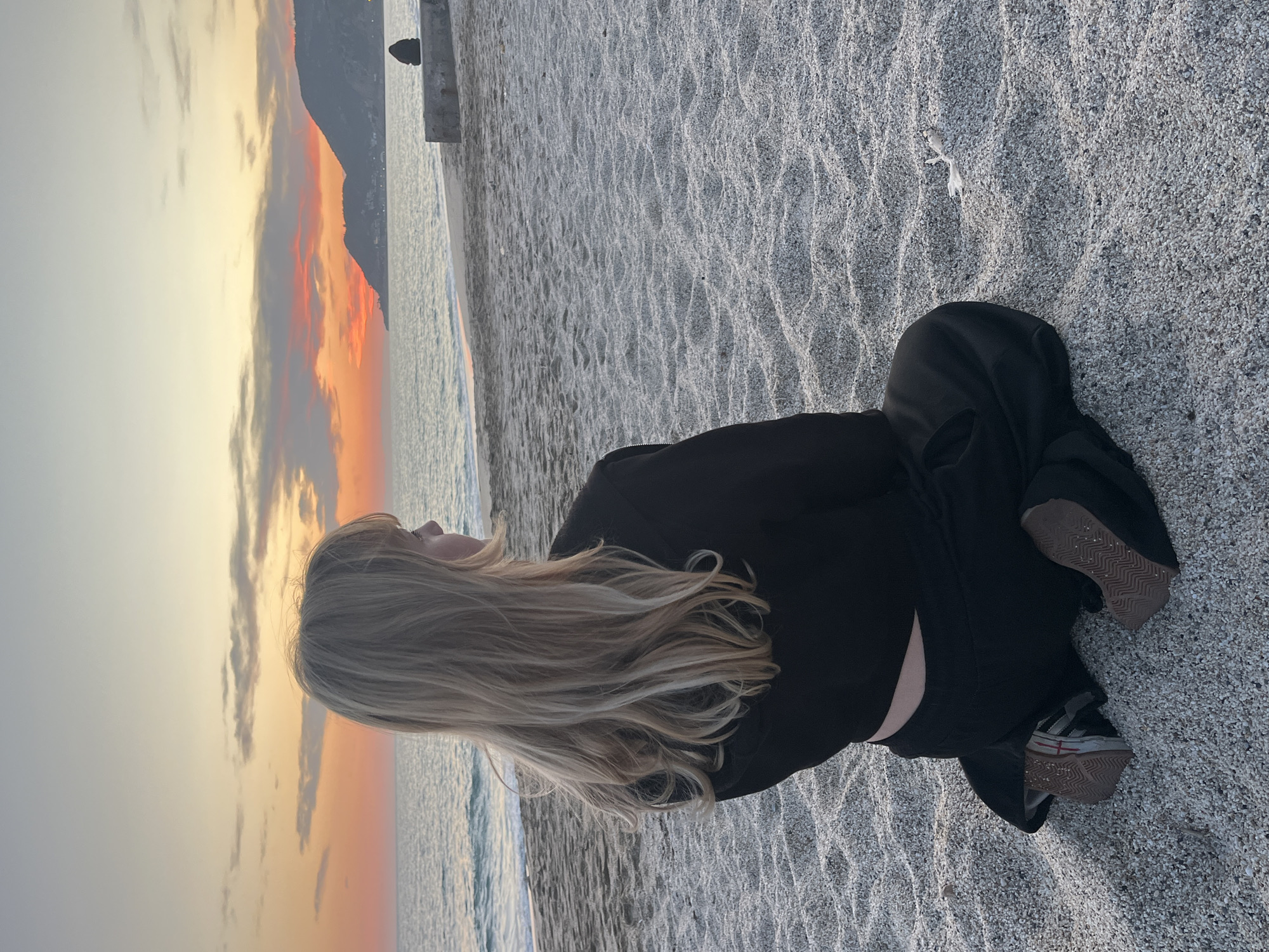 An outdoor, eye-level photograph captures a person sitting on a pebble beach at sunset in Alanya, Turkey. The primary subject is a young woman, seen from behind, with long, wavy blonde hair cascading down her back. She is dressed in a black long-sleeved top and black pants, with sneakers featuring light-colored uppers and gum soles visible on her feet.
She is seated on the light-colored, finely pebbled or coarse sand, facing the ocean and a vivid sunset. The sky is awash with warm tones of orange, pink, and yellow, blending into lighter shades and interspersed with soft, scattered clouds. The calm ocean stretches out before her, reflecting the sky's gentle glow. In the distance to the left, dark mountains create a dramatic silhouette against the horizon. A small, dark structure, possibly a pier or building, is partially visible on the right edge of the water. The beach surface shows subtle undulations. The overall atmosphere is one of serene contemplation, capturing a peaceful moment at dusk by the sea. No text is visible in the image.