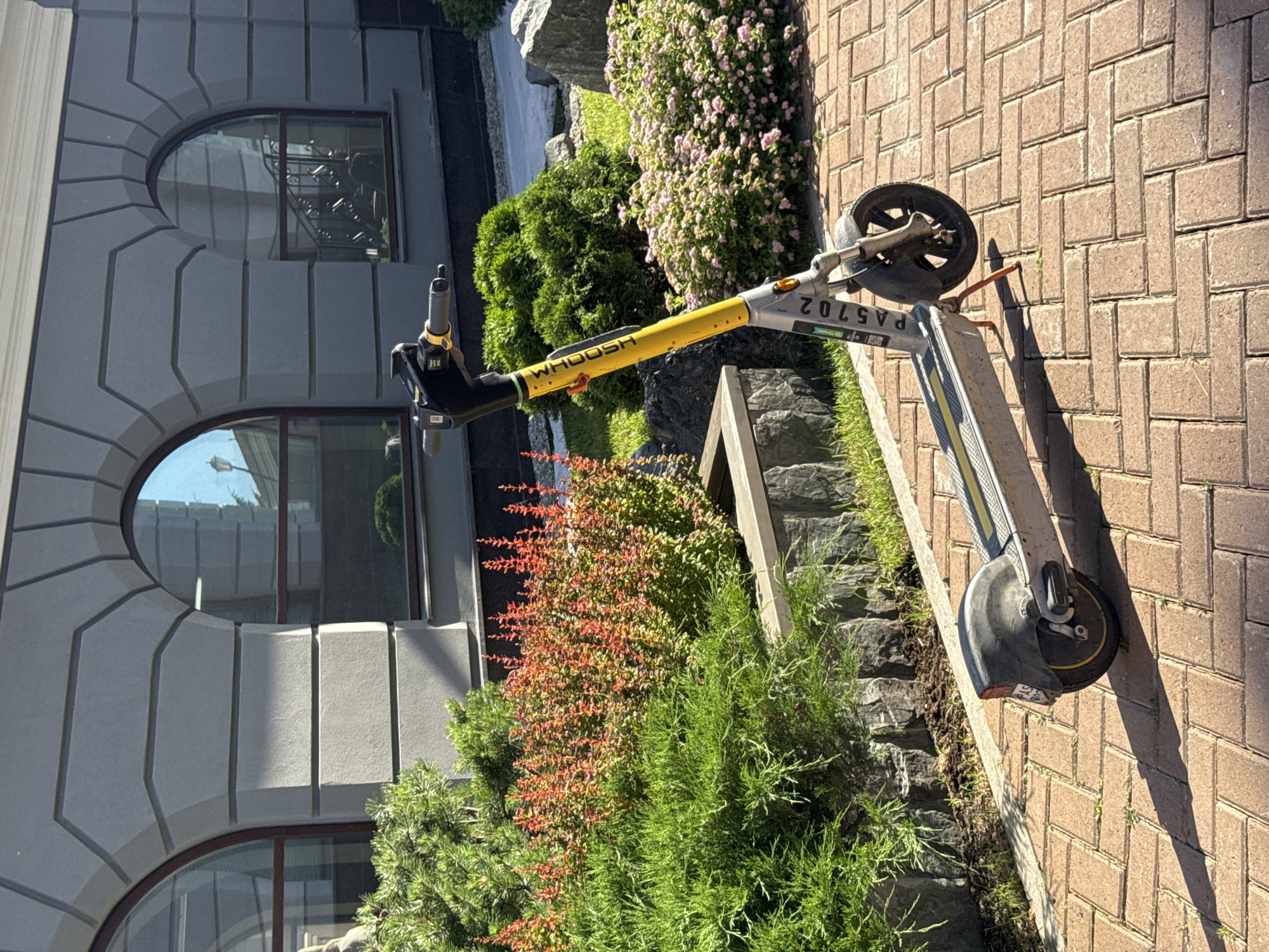 A yellow and grey electric scooter with the branding "WHOOSH" and the identification number "PA5702" is parked on a red brick pathway in front of a modern building with arched windows. The scooter has a kickstand. To the left of the scooter, there are green bushes and a hedge with red leaves. To the right, there are smaller pink flowers and large grey rocks. The building has grey, textured walls. The scene appears to be outdoors in a city setting, possibly in Yekaterinburg, Russia, given the context of the request, though no specific geographical markers are visible in the image itself. The lighting suggests it is daytime, with shadows cast by the scooter and surrounding elements. There are no people visible in the image.