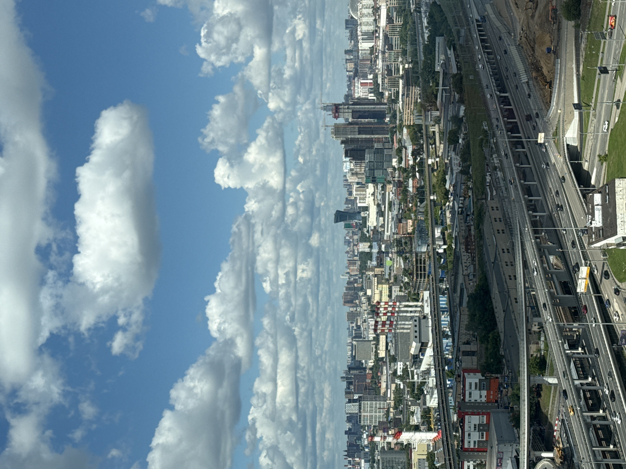 The image depicts a cityscape in Khimki, Russia, dominated by a complex network of elevated highways and urban development under a partly cloudy blue sky. The foreground features multiple levels of overpasses with active traffic, showing cars and trucks moving along the roadways. Buildings of various sizes and architectural styles are visible in the midground and background, including residential complexes, industrial facilities with smokestacks, and modern office buildings. Construction cranes suggest ongoing development in the area. The sky is filled with numerous white, fluffy clouds scattered across a vibrant blue expanse. The overall scene conveys a sense of a busy, developing urban environment.