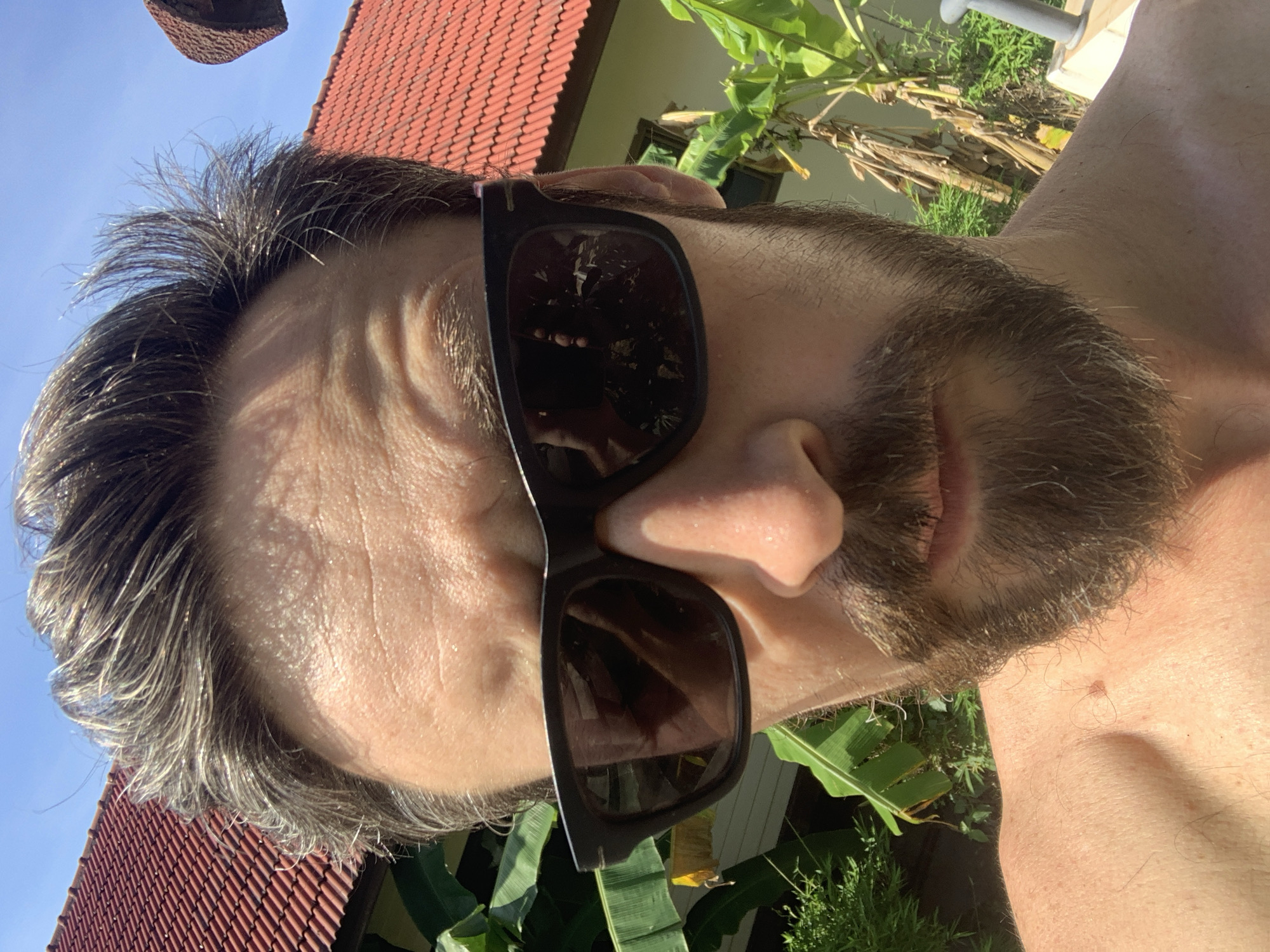 A close-up, tilted self-portrait photograph shows a light-skinned male with graying hair, stubble, and a beard. He wears dark sunglasses reflecting what appears to be a building with a red tiled roof and vegetation. The background includes a red tiled roof and green vegetation, suggestive of a tropical environment. Based on the presence of tiled roofs and lush vegetation, the location is likely in บ้านบางรักษ์, Thailand. The person is outdoors in daylight, given the natural light.