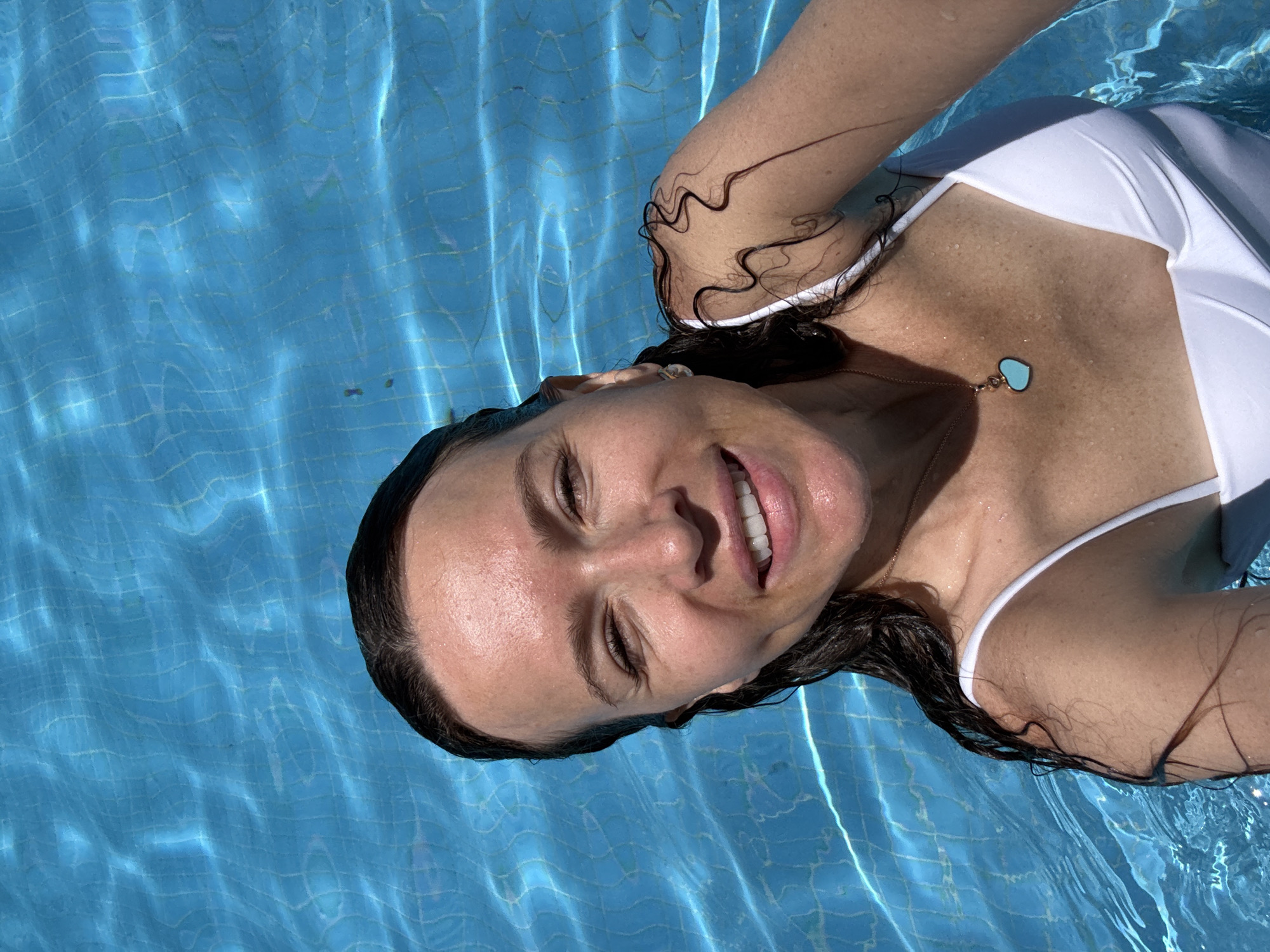 A woman with wet, dark hair is in a swimming pool. She is wearing a white swimsuit and a light blue heart-shaped necklace. Her skin is glistening with water, and she is smiling at the camera. The water in the pool is a bright blue color with a tiled bottom visible through the water. The image appears to have been taken from above, looking down at the woman. The scene is consistent with a sunny day at a resort or private pool in Dubai, United Arab Emirates.