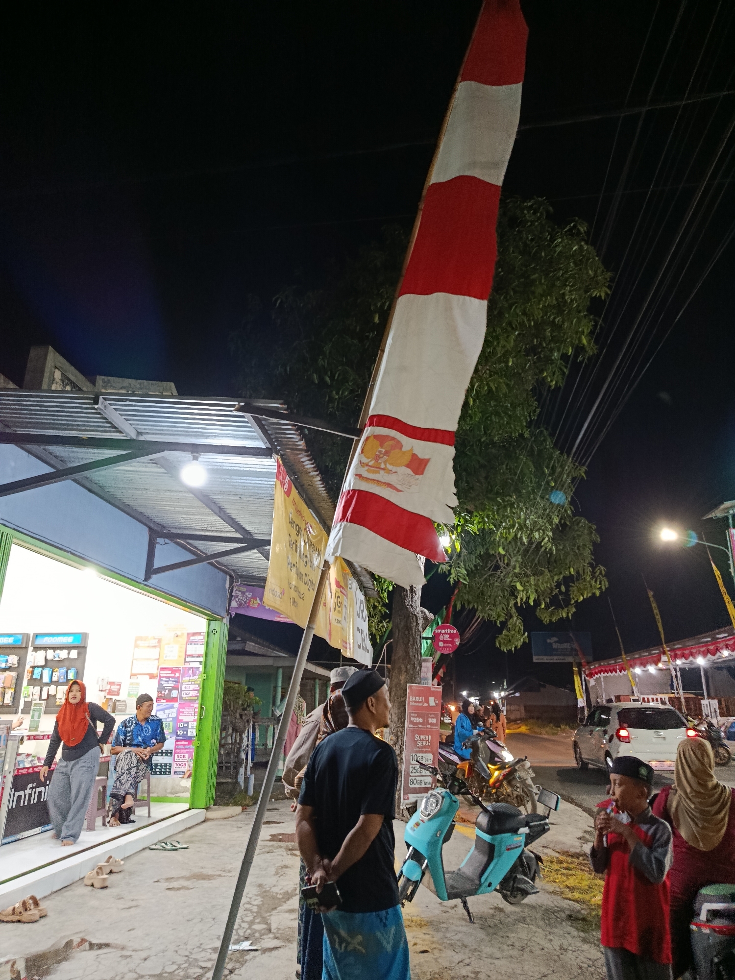 In the city of Ramban Wetan, Indonesia, at night, a scene unfolds outside a shop with a corrugated metal roof. A large Indonesian flag, red and white, is prominently displayed, attached to a pole. The flag bears an emblem. Several individuals are present. On the left, a woman in a red hijab and grey pants stands near the shop entrance, which is brightly lit and displays various products. Two men are seated inside the shop, one in a blue patterned shirt and the other in a black shirt and cap, holding a phone. To the right, a boy in a red shirt and black cap is eating something from a wrapper. Behind him, a woman in a beige hijab is visible. A turquoise scooter is parked in the foreground, and a white car is in the background. Trees and power lines are visible, and streetlights illuminate the area. The ground is paved and wet, suggesting recent rain. The overall impression is a bustling street scene at night.