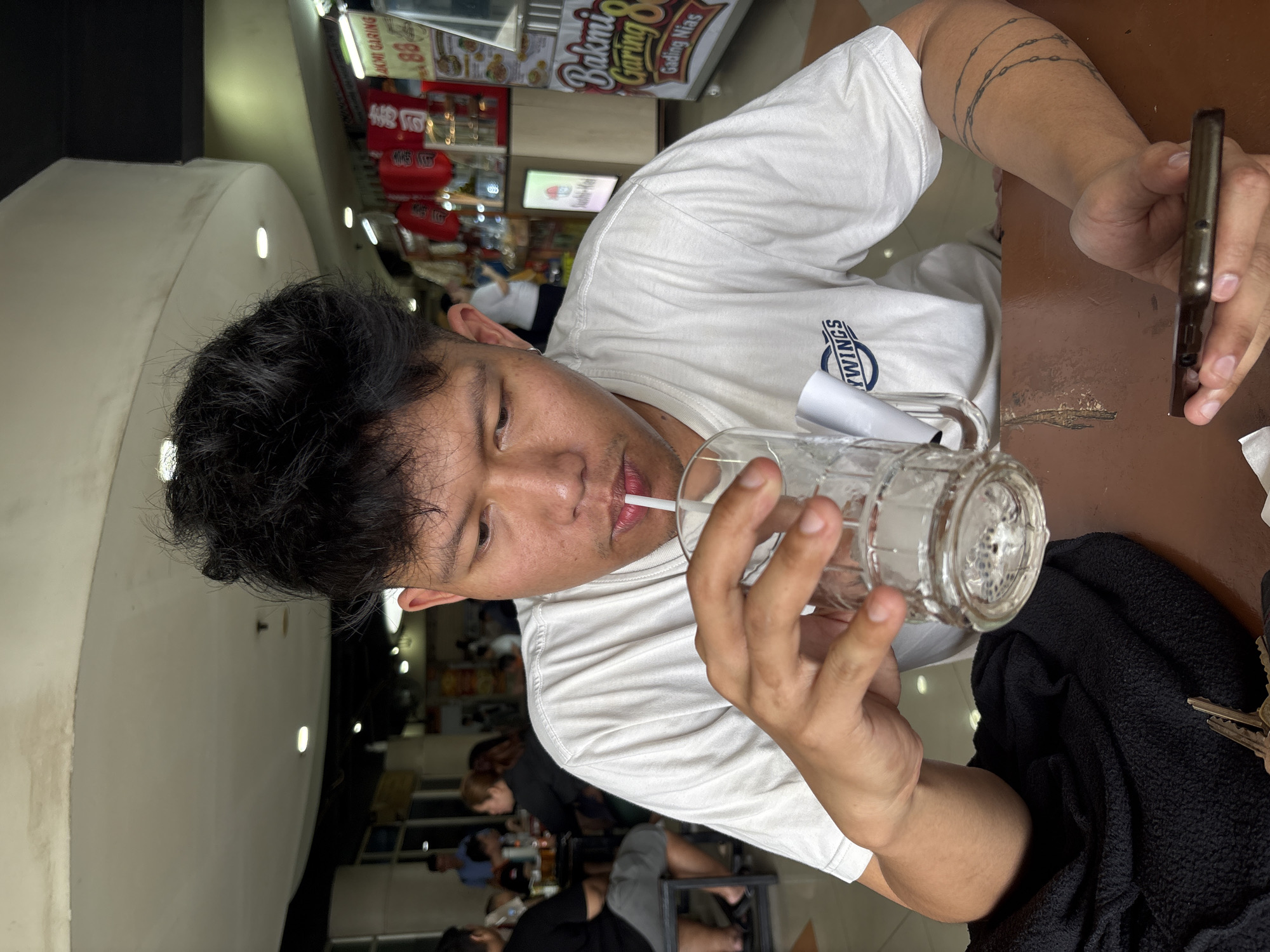 A young man with dark, spiky hair is seated at a table, drinking from a glass with a straw. He is wearing a white t-shirt with a circular logo on the left chest that reads "MYWINGS." His right arm, visible from the elbow down, has a tattoo of interconnected circular chains. He is holding a smartphone in his left hand. In the background, there are other people seated at tables and food stalls with red lanterns and signs, suggesting a food court or market setting. One sign prominently displays "Bakmi Garing 88 Gading Nias," indicating the location is likely in the Gading Nias area of North Jakarta, Indonesia. The environment appears to be an indoor public space, possibly a mall food court, with a white, curved ceiling and bright lighting.