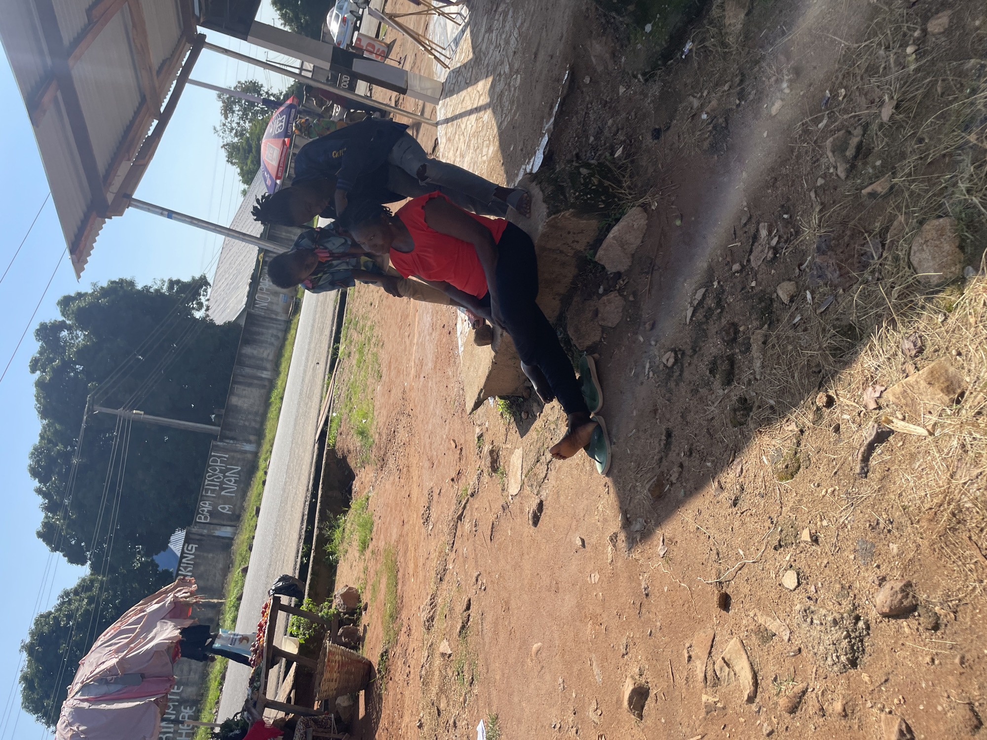 In Jos, Nigeria, a woman in a red top and black pants sits on a concrete ledge outdoors, looking to her left. Two other individuals, a young person in a patterned shirt and another in a dark jacket and ripped jeans, stand behind her. The scene unfolds on a dirt-covered area next to a road with a concrete drain. To the left, a market stall displays some produce under a large, draped umbrella. In the background, a wall with graffiti reads "KING BAA FITSPARI A NAN." Further in the distance, trees and utility poles are visible under a clear blue sky. The area appears to be a roadside market or gathering spot.