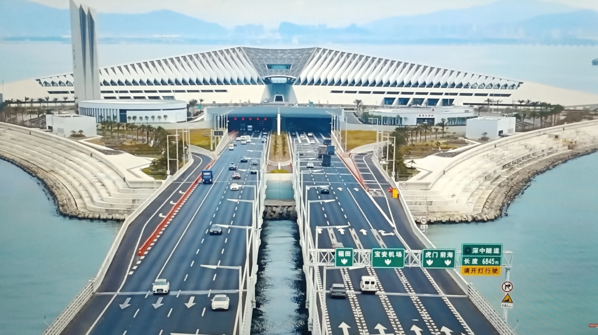 This image captures a wide, multi-lane elevated highway curving towards a modern tunnel entrance. The highway is bordered by water on both sides, with concrete barriers and white railings. Numerous vehicles, including cars and trucks, are traveling on the highway. In the background, a large, architecturally striking building with a distinctive angled roofline stands prominently, overlooking the water and the highway. The sky is overcast with a pale blue hue. The image appears to be a daytime aerial or high-angle shot of a contemporary infrastructure project. The location is not in Dubăsari town council, Moldova, as this appears to be a modern coastal infrastructure project likely in East Asia.
