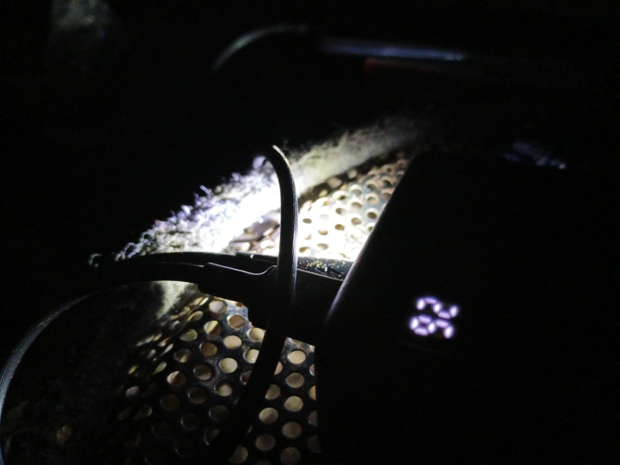 The image shows a close-up, low-light view of what appears to be a piece of electronic equipment. In the foreground, a black device with a digital display showing the number "26" is partially visible. The display is illuminated with a purple glow. To the left of this device, a curved black metal rod or cable is suspended over a circular, perforated metal surface. This perforated surface is lit from below, revealing a pattern of evenly spaced circular holes. The background is dark and indistinct, with some fuzzy, bright areas suggesting light sources or reflections. The overall scene is dark, with the main subjects illuminated by focused light. There are no people or identifiable urban features present.
