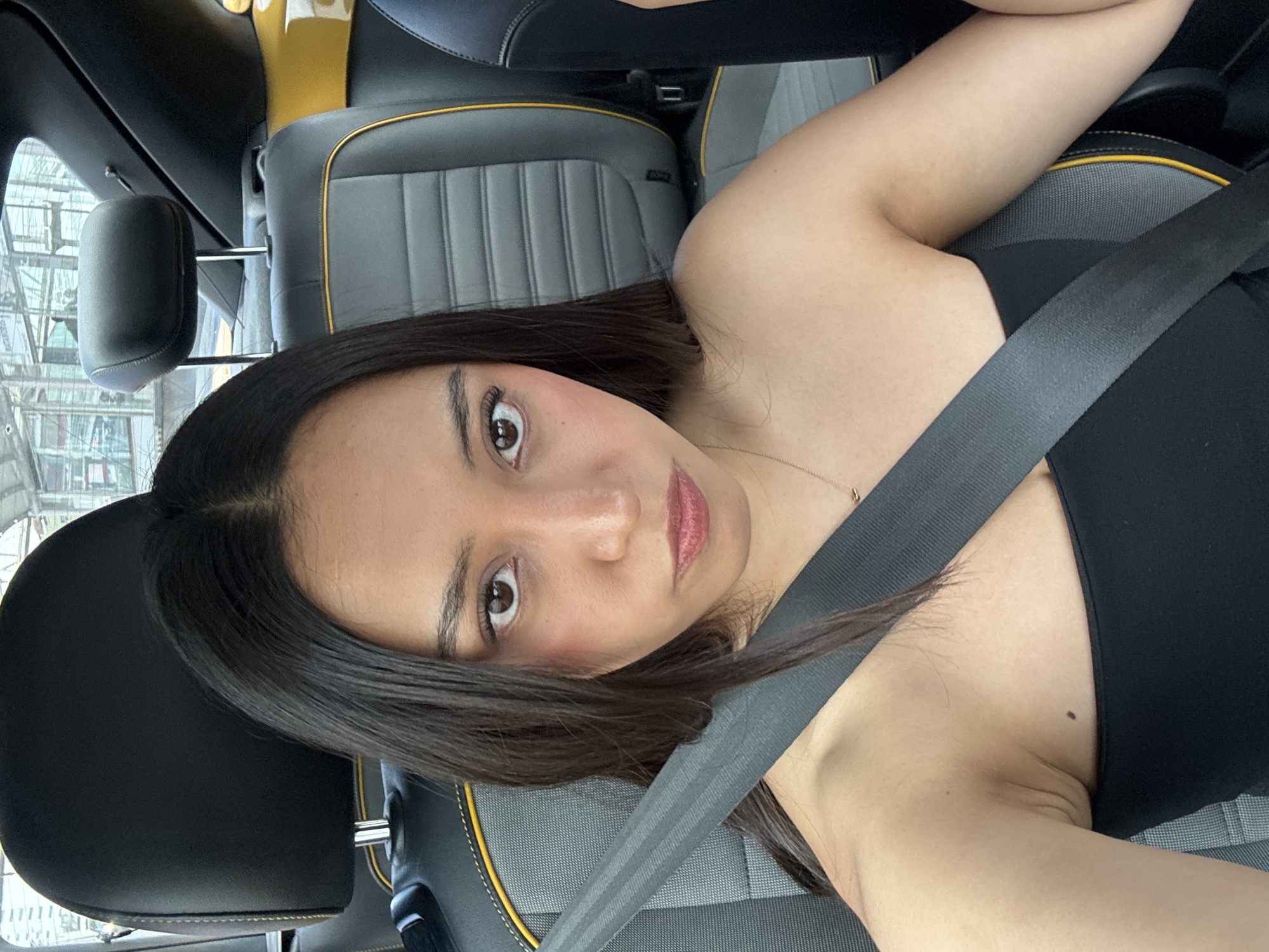 A young woman with brown hair and light skin is seated in the driver's seat of a car, wearing a black top and a seatbelt. She is looking at the camera with brown eyes and a slightly smiling expression. The car has gray leather seats with yellow piping. Outside the car window, a blurry view of a building suggests an urban environment. The location is Puebla, Mexico.
