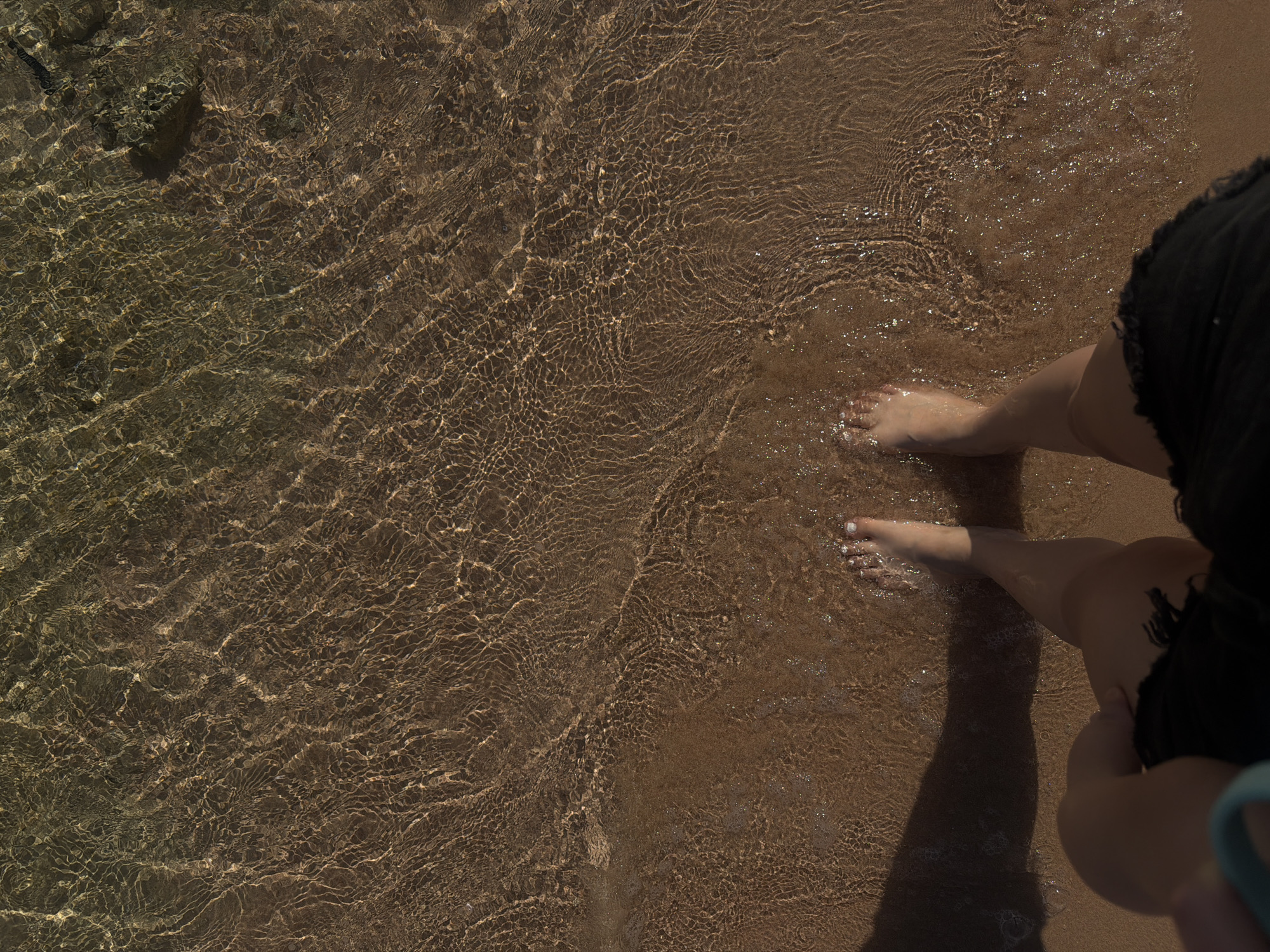 The image depicts a top-down view of a person's bare feet standing in shallow, clear water at the edge of a sandy beach. The water is illuminated by sunlight, creating rippling patterns on the seabed, which appears to be composed of sand and some small rocks. The feet are positioned in the lower center of the frame, with one foot slightly ahead of the other. The person is wearing dark shorts, and a shadow is cast to the left, suggesting the sun is to the right. The scene is characteristic of a beach environment, likely in Sharm El Sheikh, Egypt, known for its clear waters and sandy shores.
